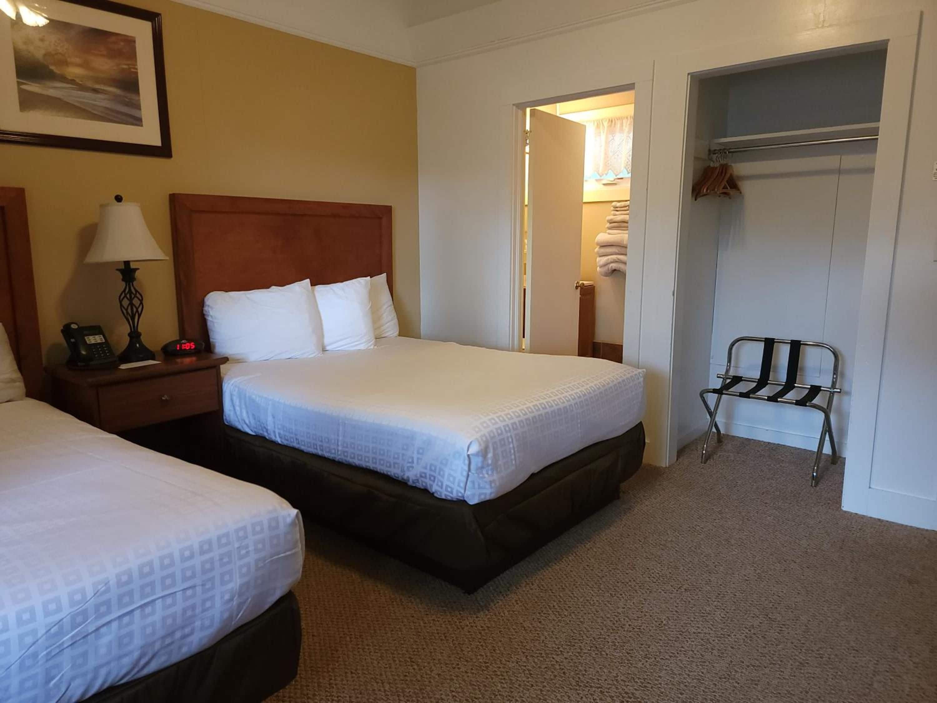 A hotel room with two beds, a nightstand with a lamp, a clock, and a coat rack.