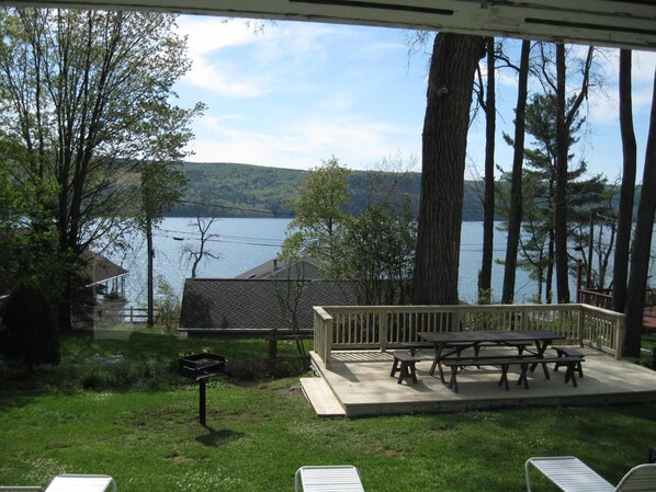 A deck with picnic tables overlooking a lake, surrounded by trees and a grassy area.