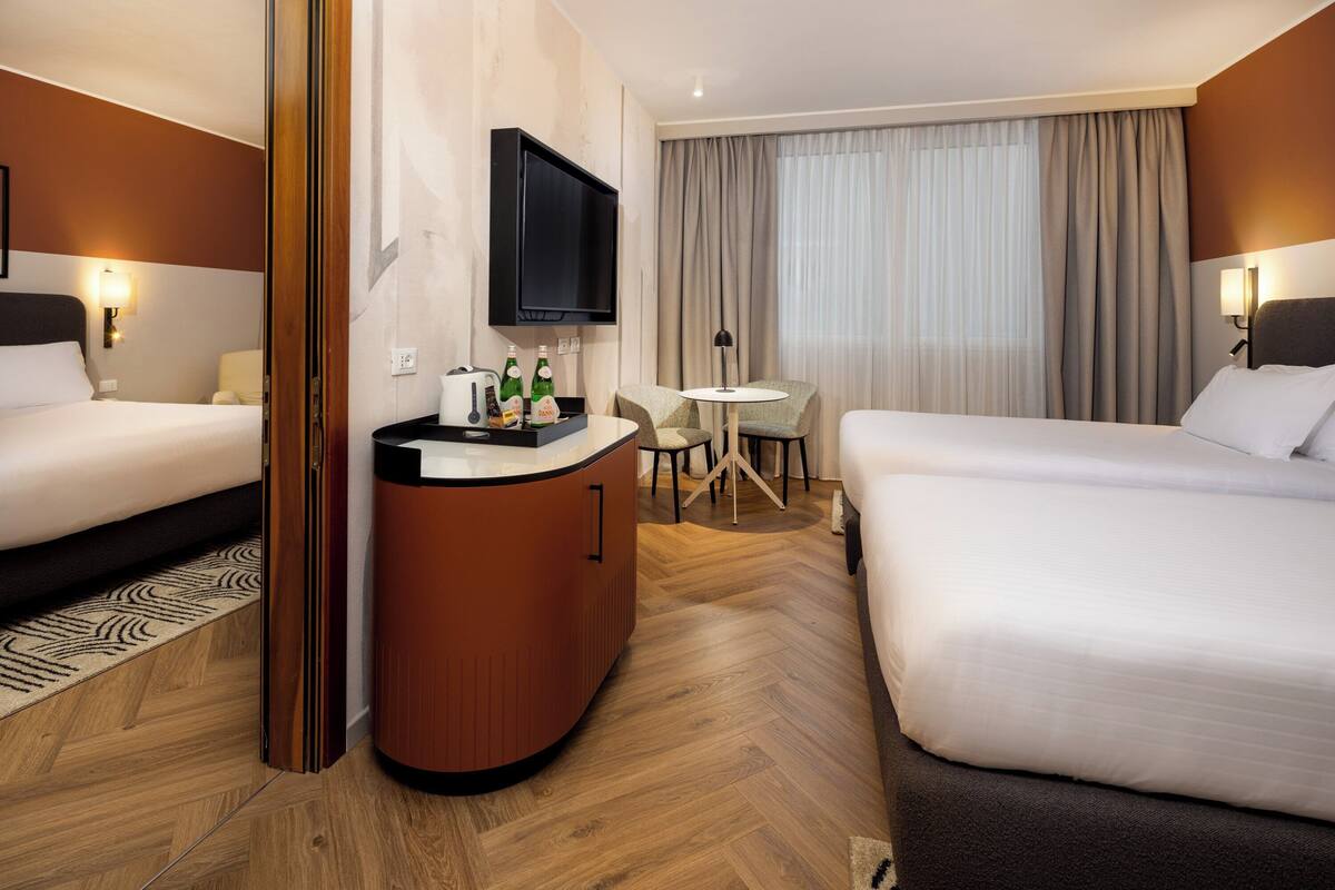 Habitación de hotel con dos camas, televisión, minibar, una pequeña mesa con sillas y un ventanal con cortinas.