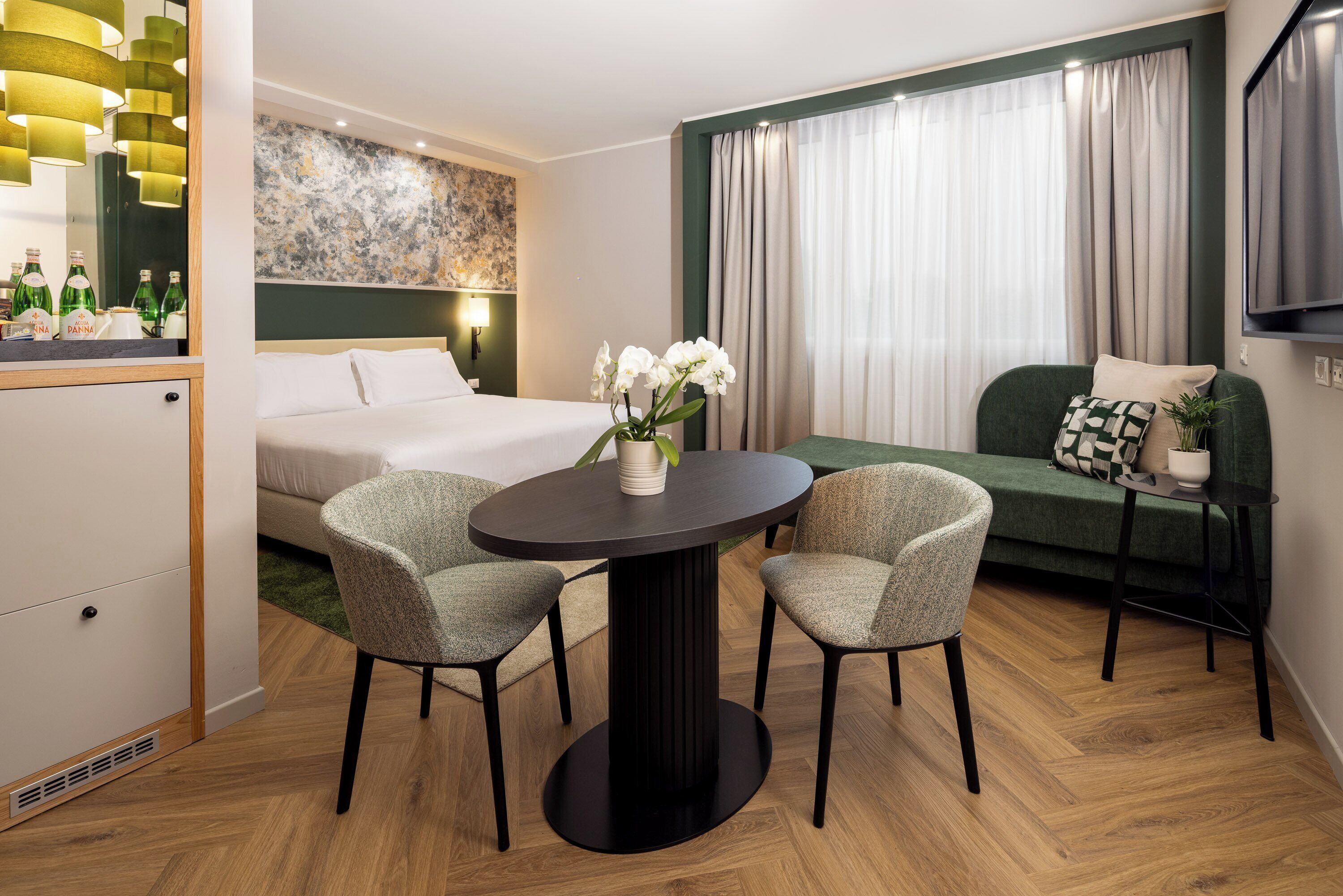 A modern hotel room with a bed, a round dining table with two chairs, a sofa, and a TV mounted on the wall.