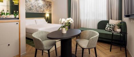 A modern hotel room with a bed, a round dining table with two chairs, a sofa, and a TV mounted on the wall.