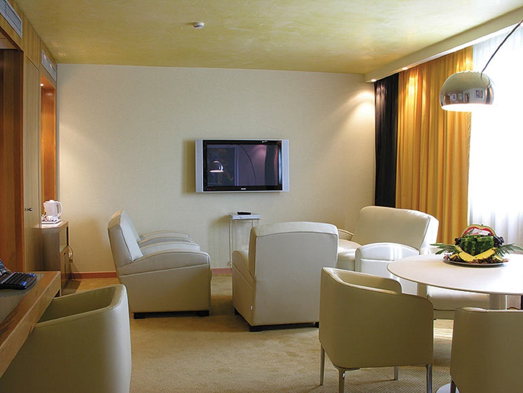 Ein Hotelzimmer mit Wandfernseher, einem kleinen Tisch mit Obstschale, zwei Sesseln und einem Schreibtisch mit Computer.