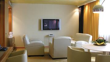 Ein Hotelzimmer mit Wandfernseher, einem kleinen Tisch mit Obstschale, zwei Sesseln und einem Schreibtisch mit Computer.
