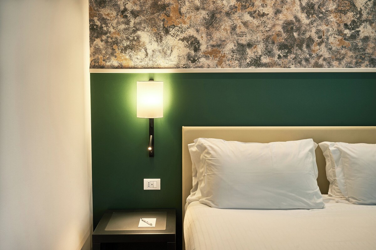 Una habitación de hotel con una pared verde, una cama blanca con dos almohadas, una lámpara de noche y una mesita de noche con un bolígrafo y bloc de notas.