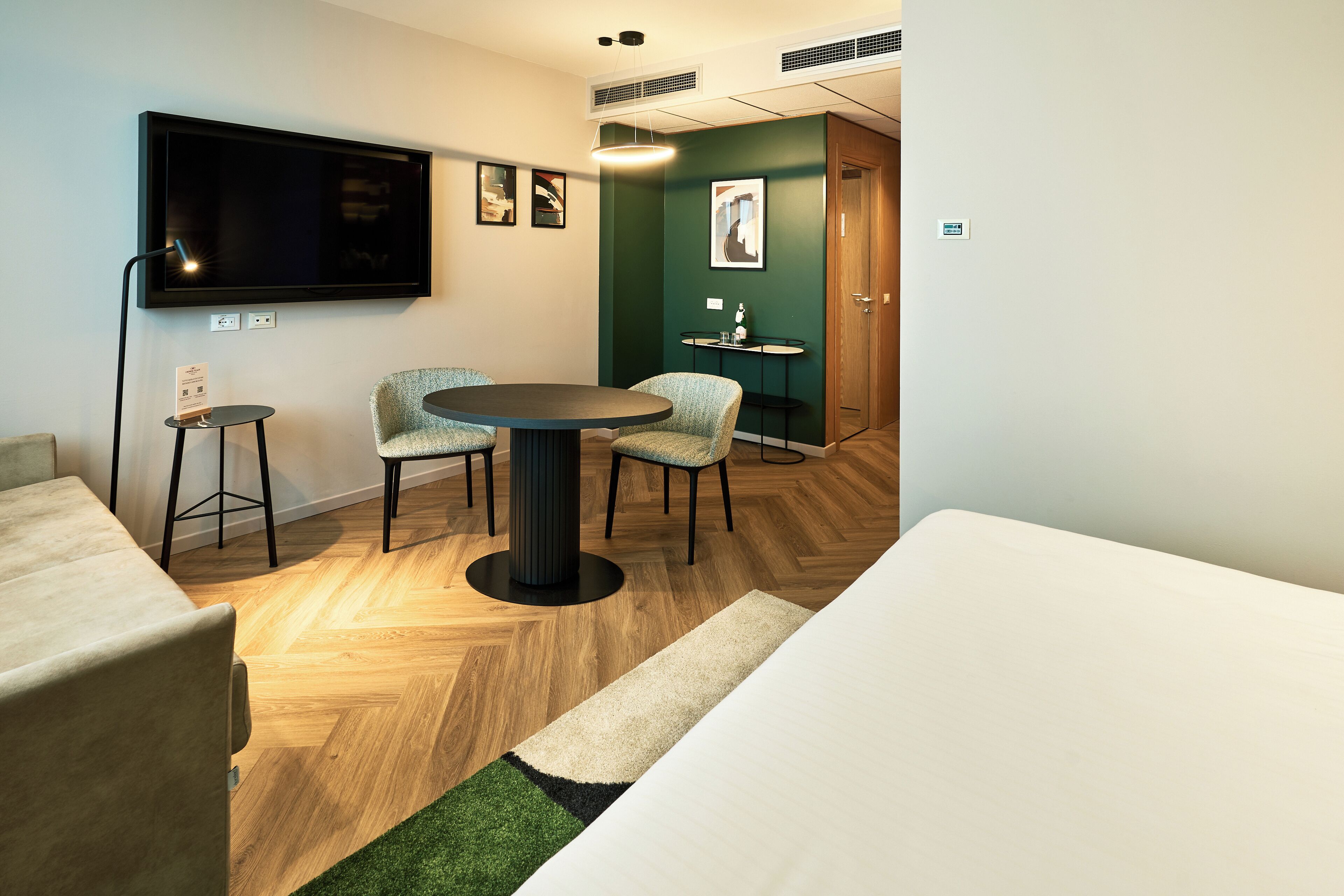 Una habitación de hotel moderna con televisor de pantalla plana, una mesa redonda, dos sillas, un sofá y una mesita auxiliar.