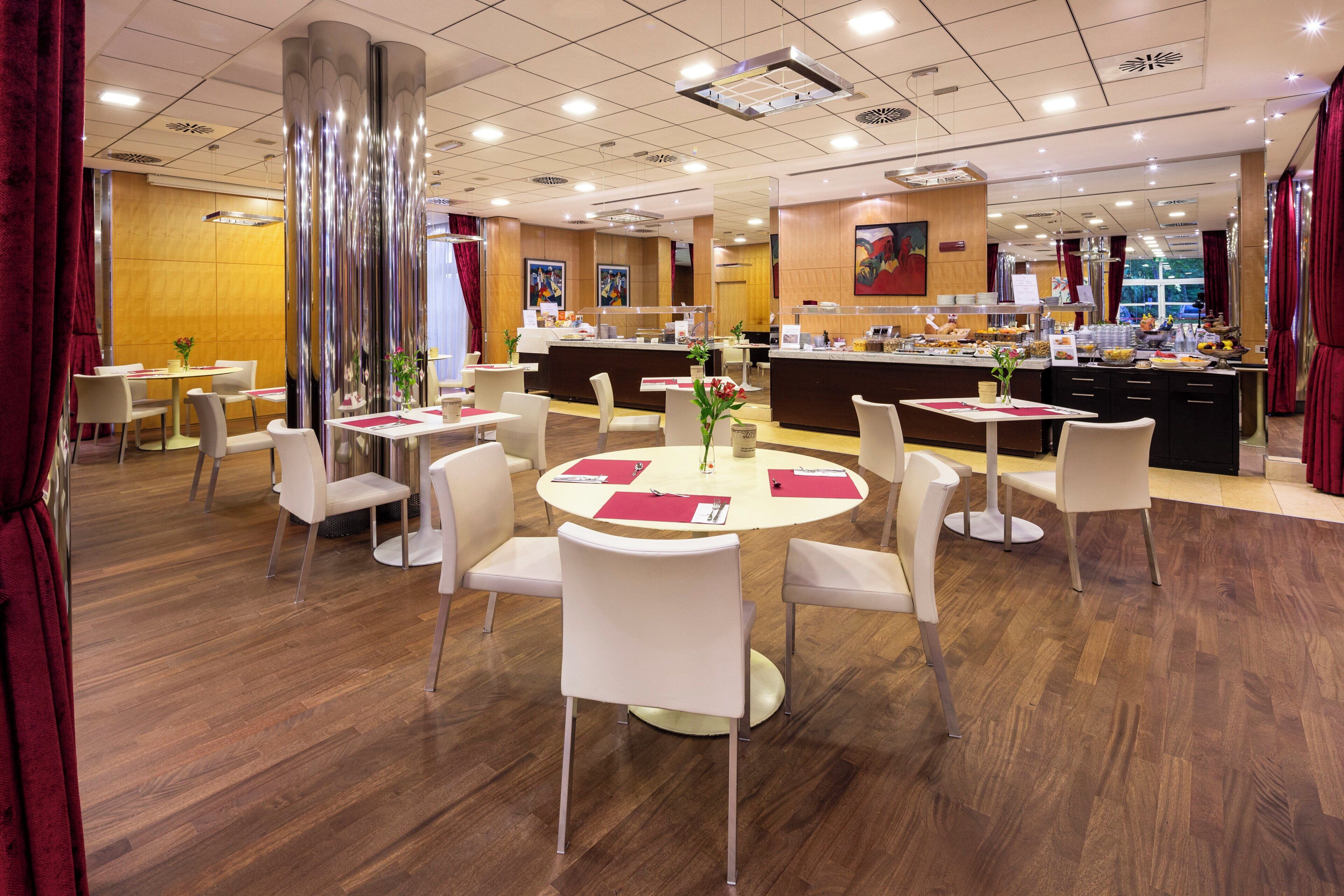 A hotel dining area with round tables, white chairs, a buffet counter, and a large mirror reflecting the space.