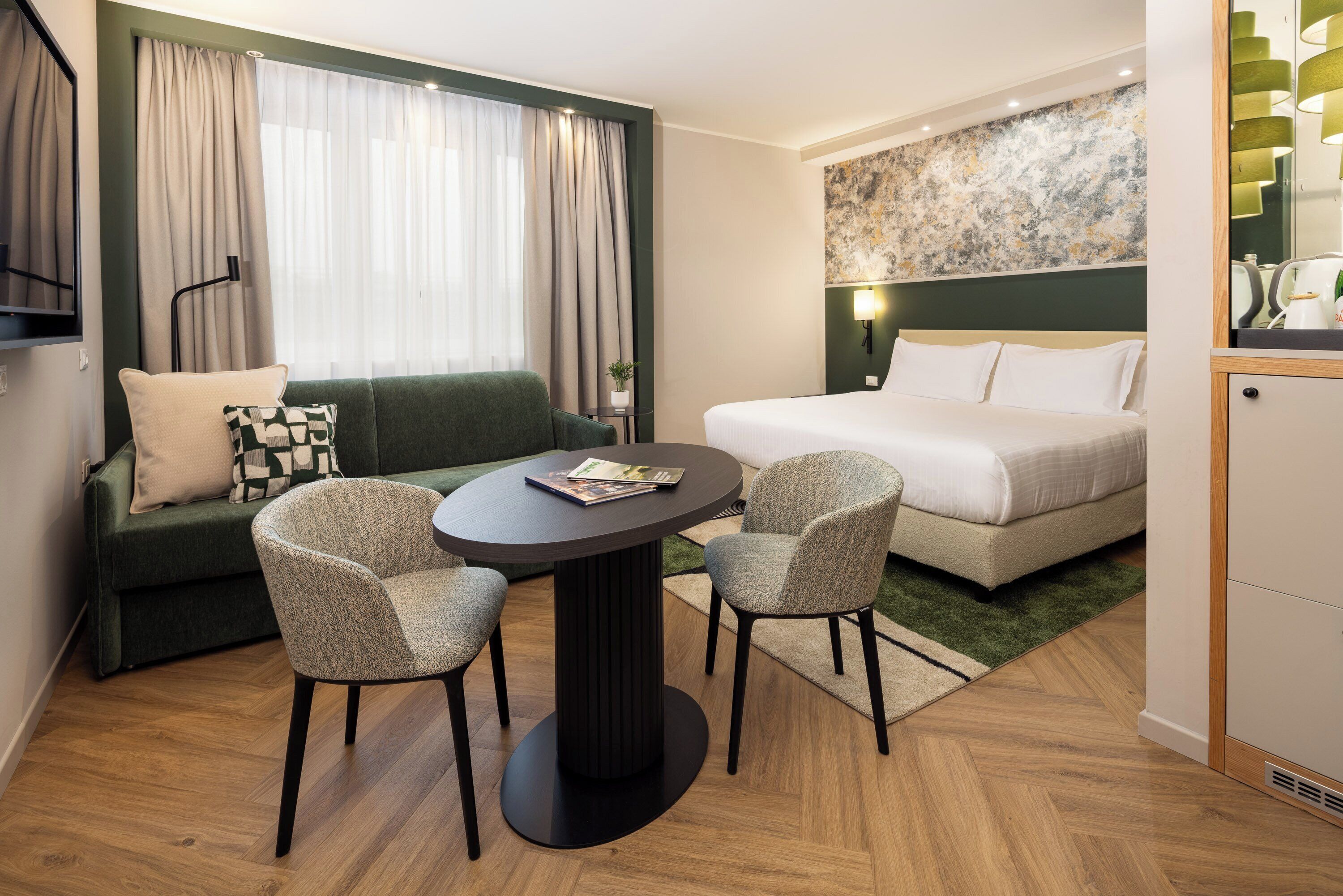 A hotel room with a bed, a green sofa, two armchairs, a round table, and a small side table.