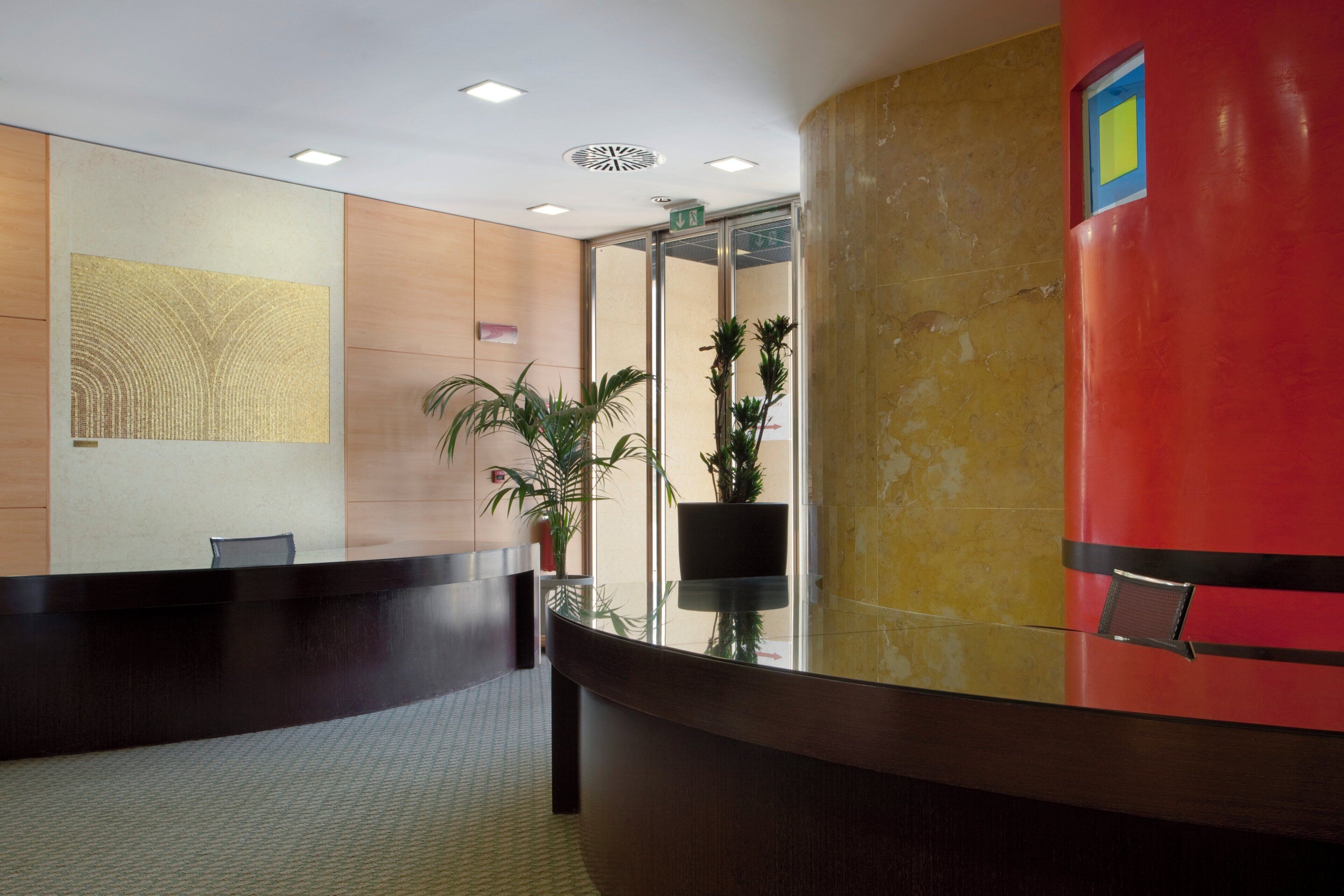 A reception area with a reception desk, a plant, and a wall with a textured artwork.