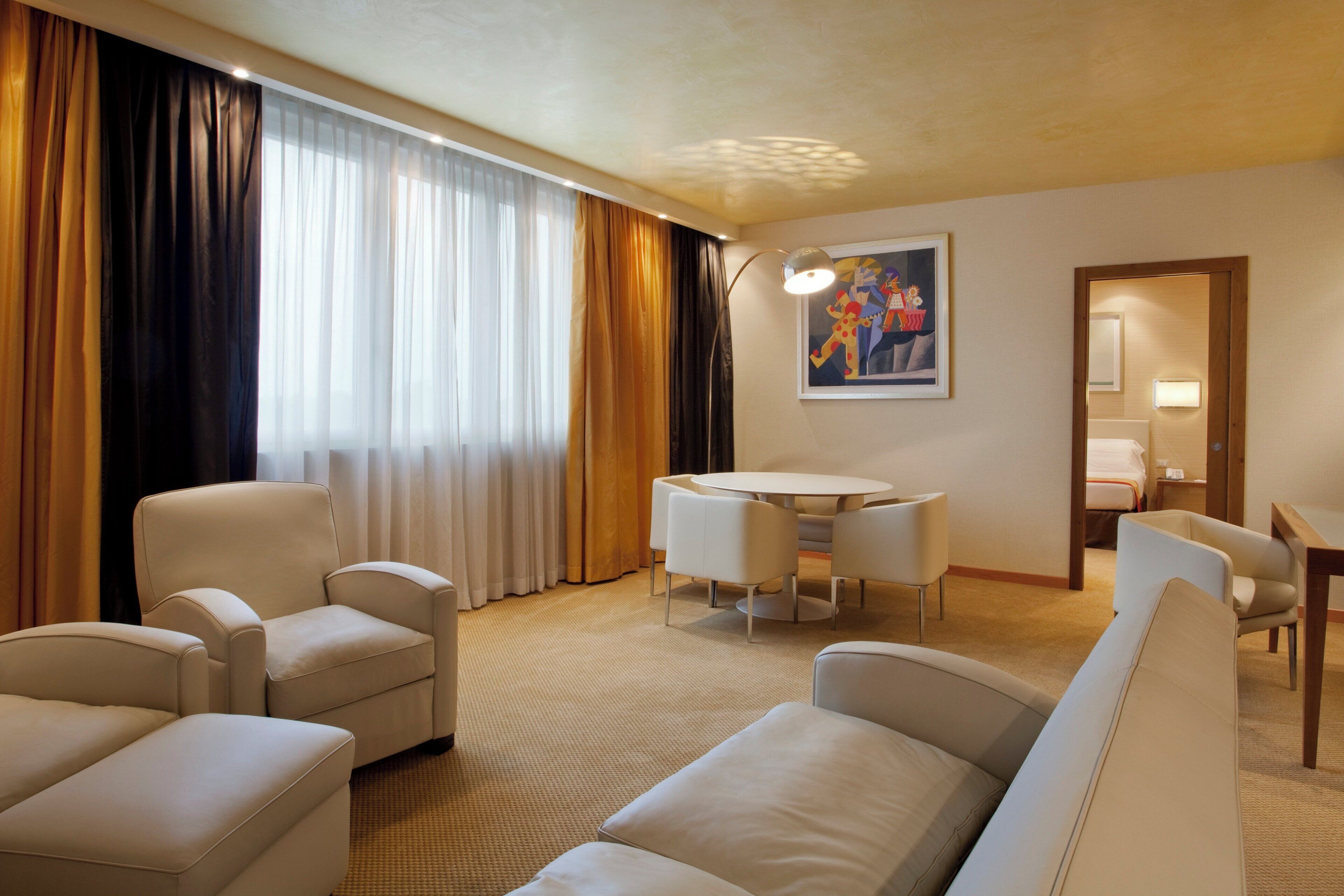 A hotel room with a seating area, a dining table, and a painting on the wall.