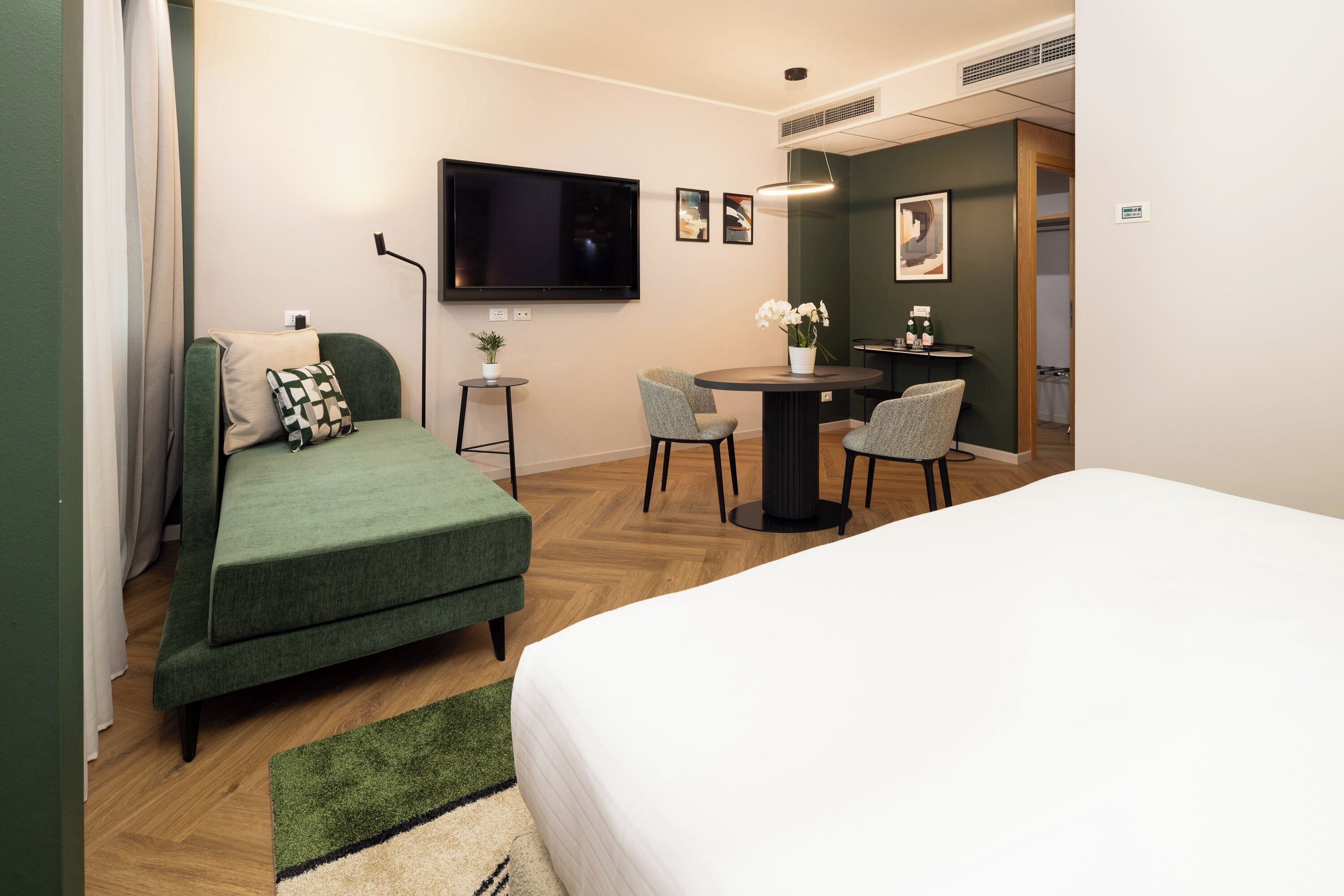 A hotel room with a green sofa, a bed, a small table with chairs, a TV mounted on the wall, and a potted plant.