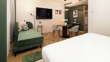 A hotel room with a green sofa, a bed, a small table with chairs, a TV mounted on the wall, and a potted plant.