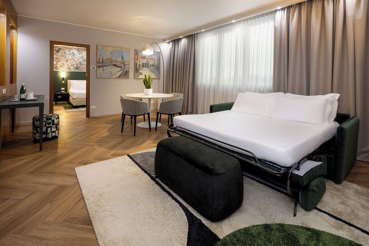 Una habitación de hotel con una cama grande, un escritorio con una silla, una mesita con una silla y un cuadro en la pared.