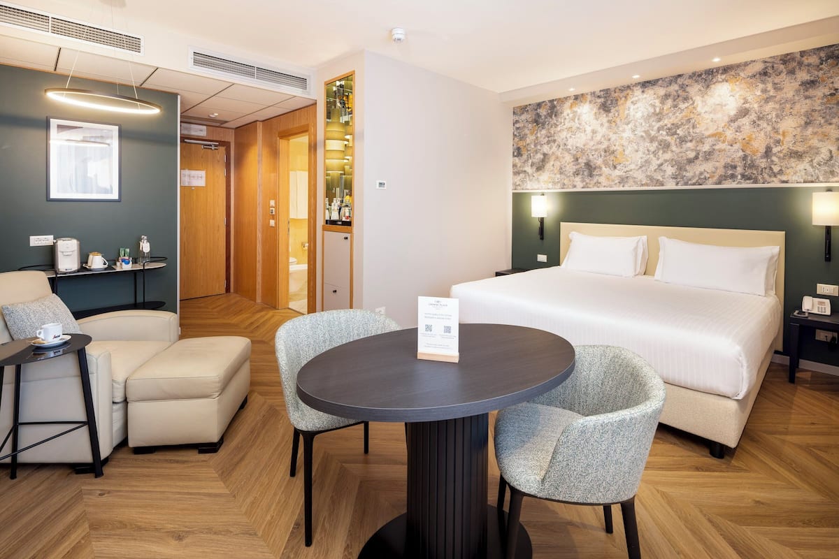 A hotel room with a bed, a round table with chairs, a small sofa, a coffee machine, and a wall-mounted artwork.