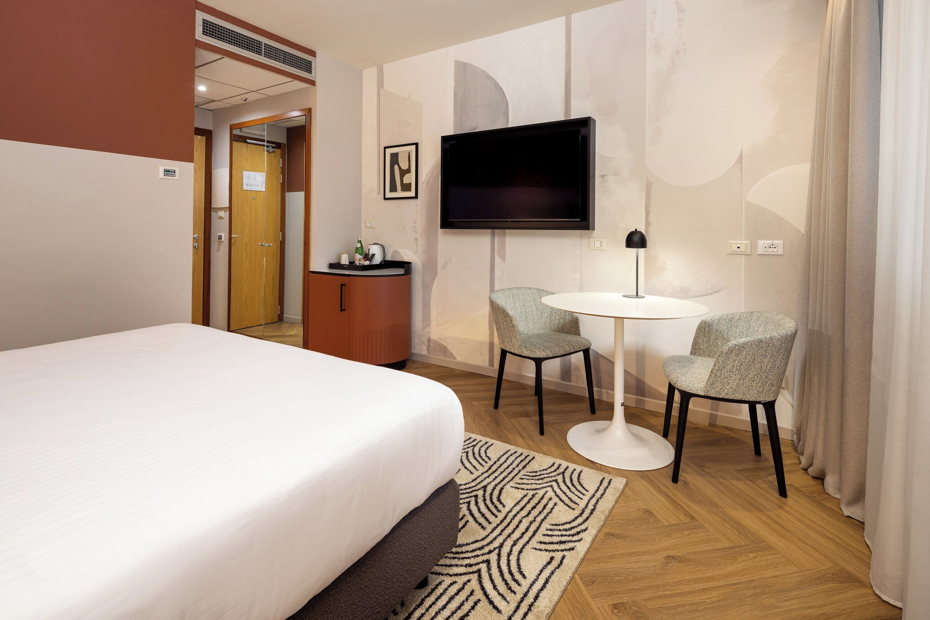 A hotel room with a bed, a TV mounted on the wall, a small desk with a chair, and a patterned rug.