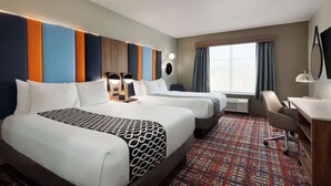 Une chambre d’hôtel avec deux lits, un bureau, une chaise, une grande fenêtre avec des rideaux et un mur muni d’un luminaire.