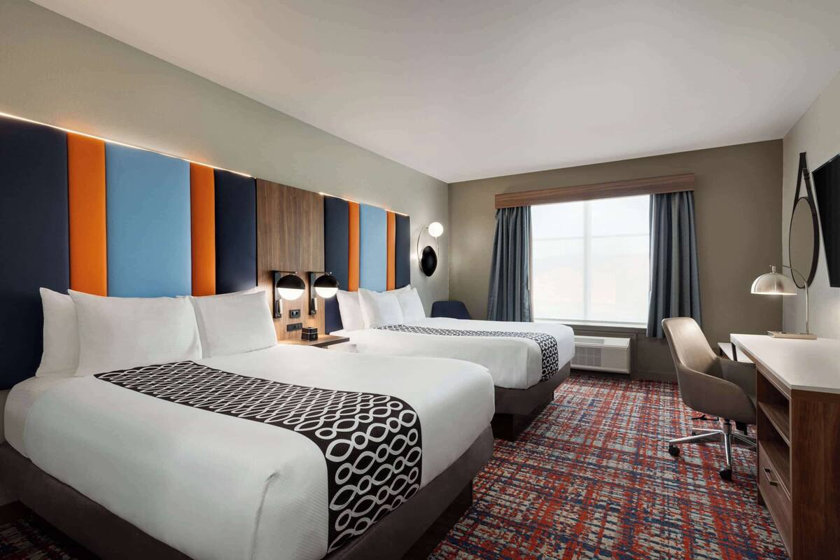 Une chambre d’hôtel avec deux lits, un bureau, une chaise, une grande fenêtre avec des rideaux et un mur muni d’un luminaire.