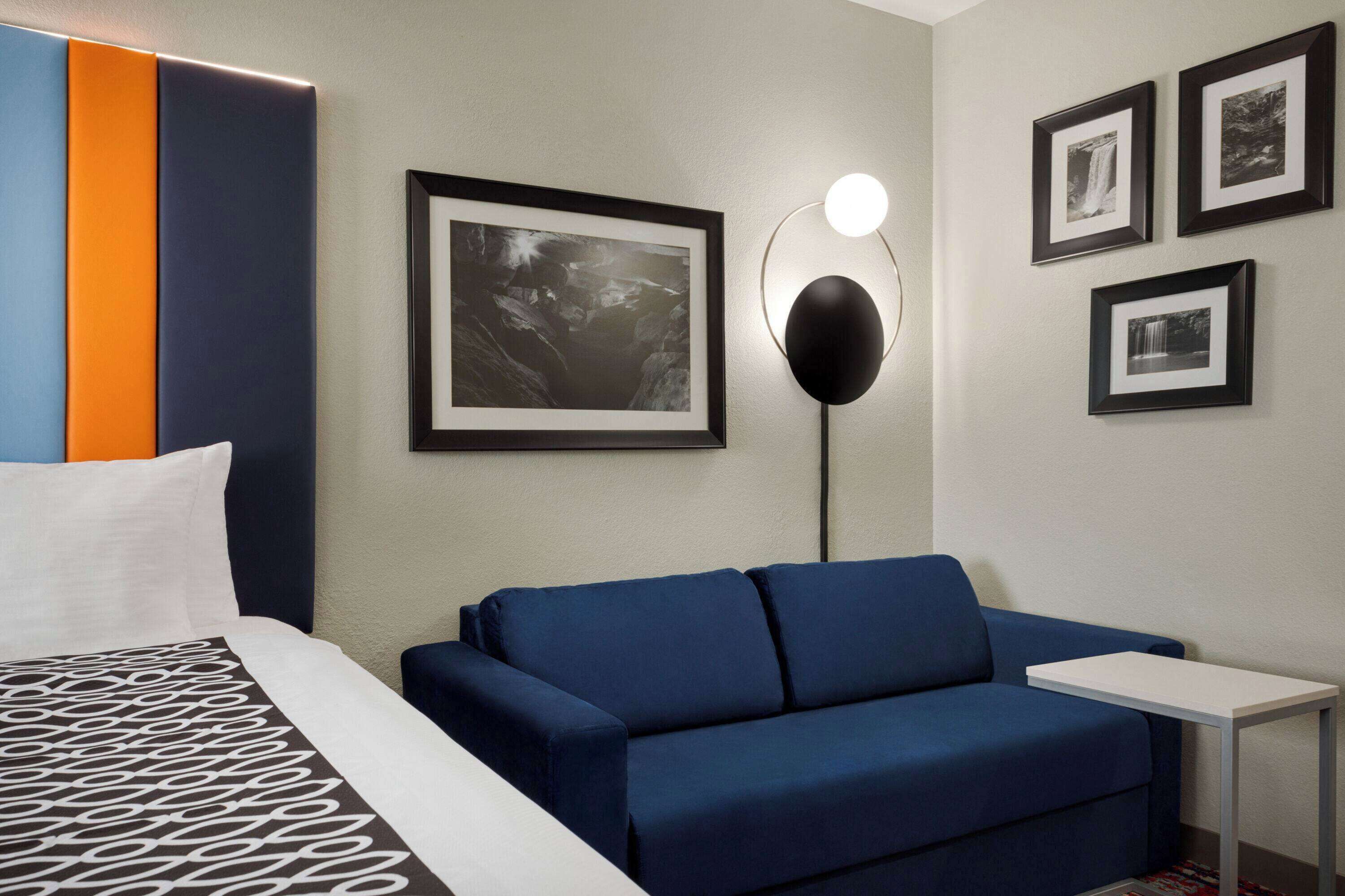 A hotel room with a bed, a blue sofa, a small white table, and three framed pictures on the wall.