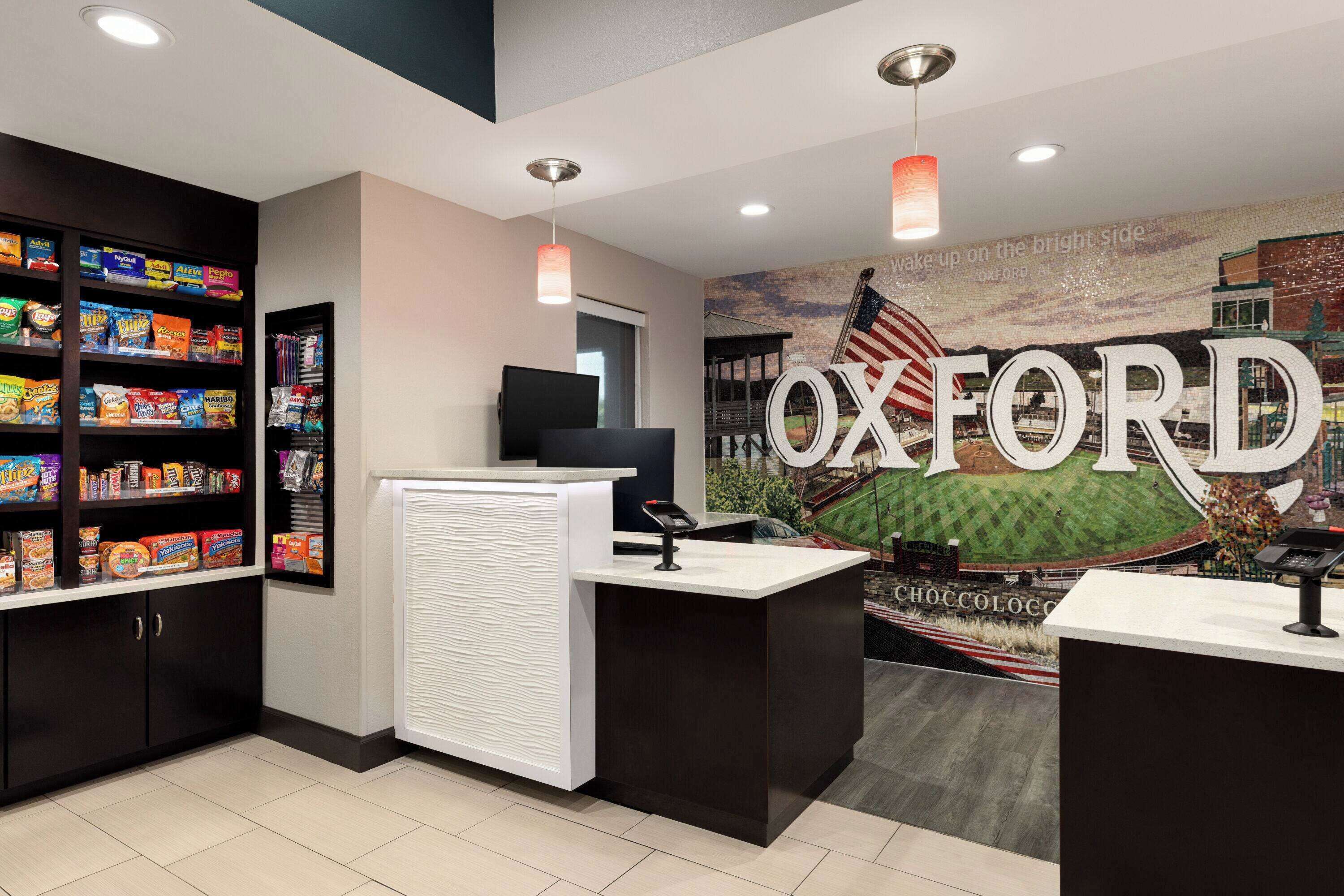 Zona de recepción con un gran mural de 'OXFORD', un estante con aperitivos y un mostrador de recepción moderno.
