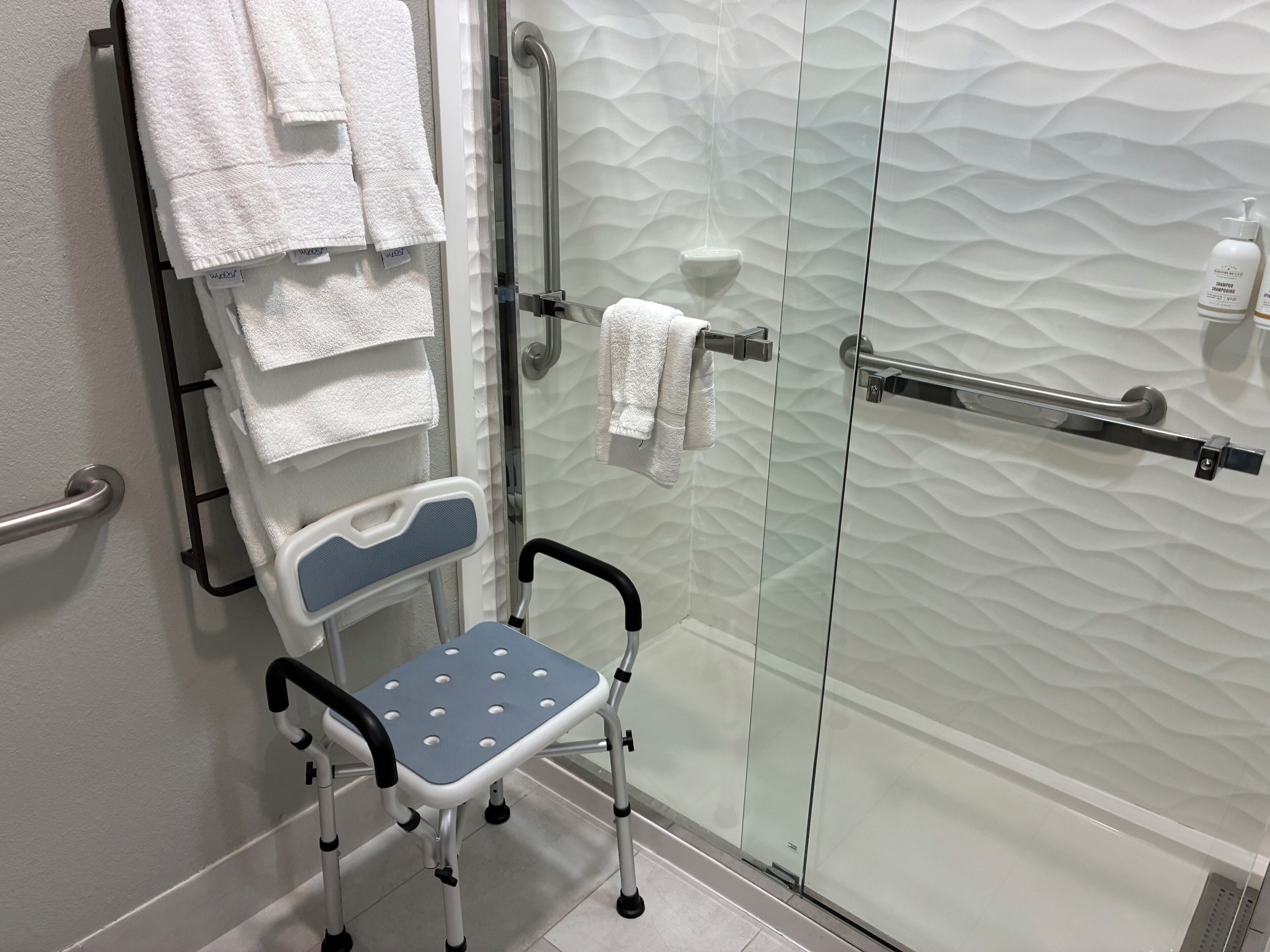Baño con ducha, puerta de vidrio, toallero con varias toallas, cabezal de ducha manual y un dispositivo de asistencia para movilidad.