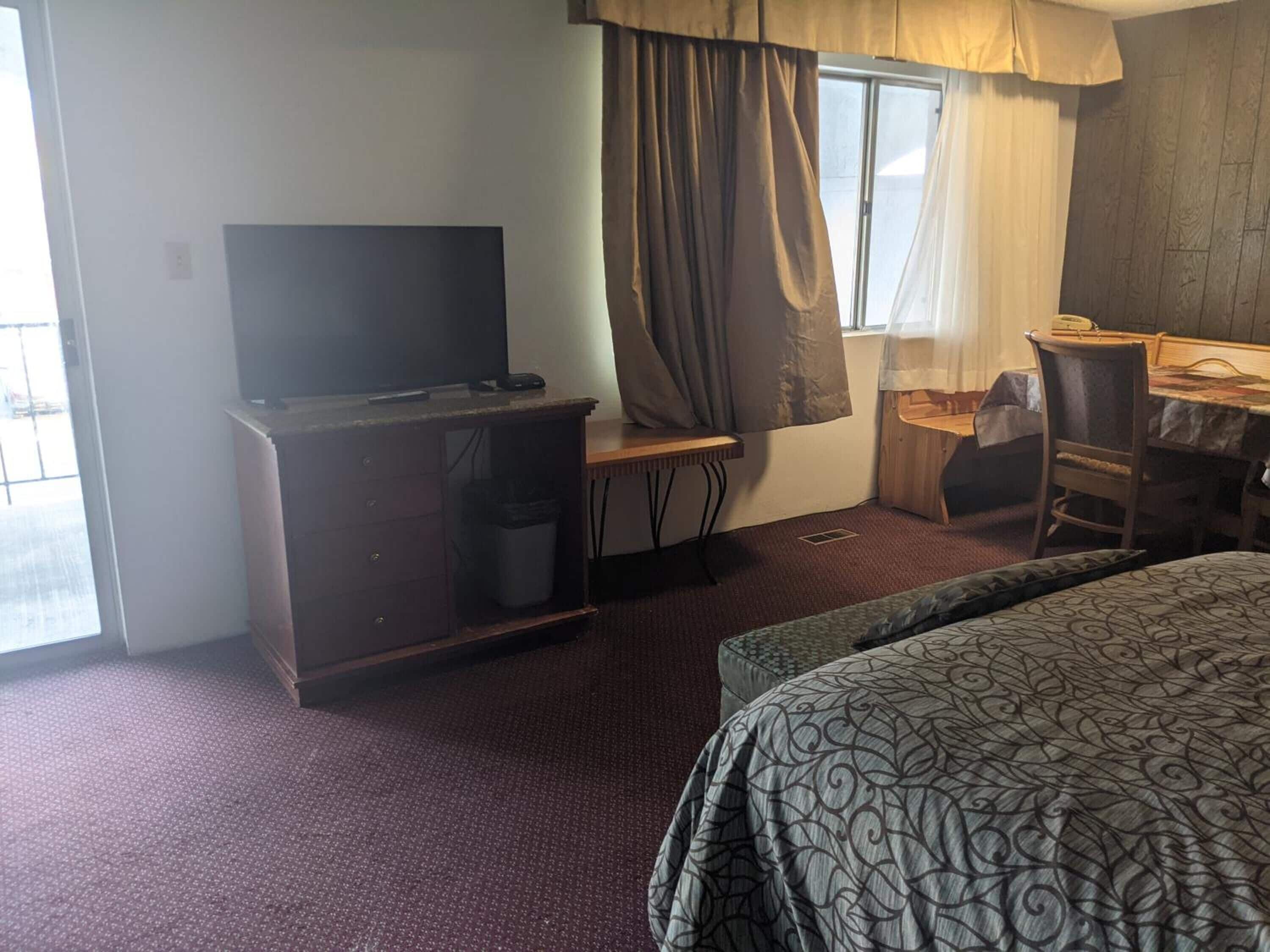 A hotel room with a bed, a desk with a chair, a television, and a window with curtains.