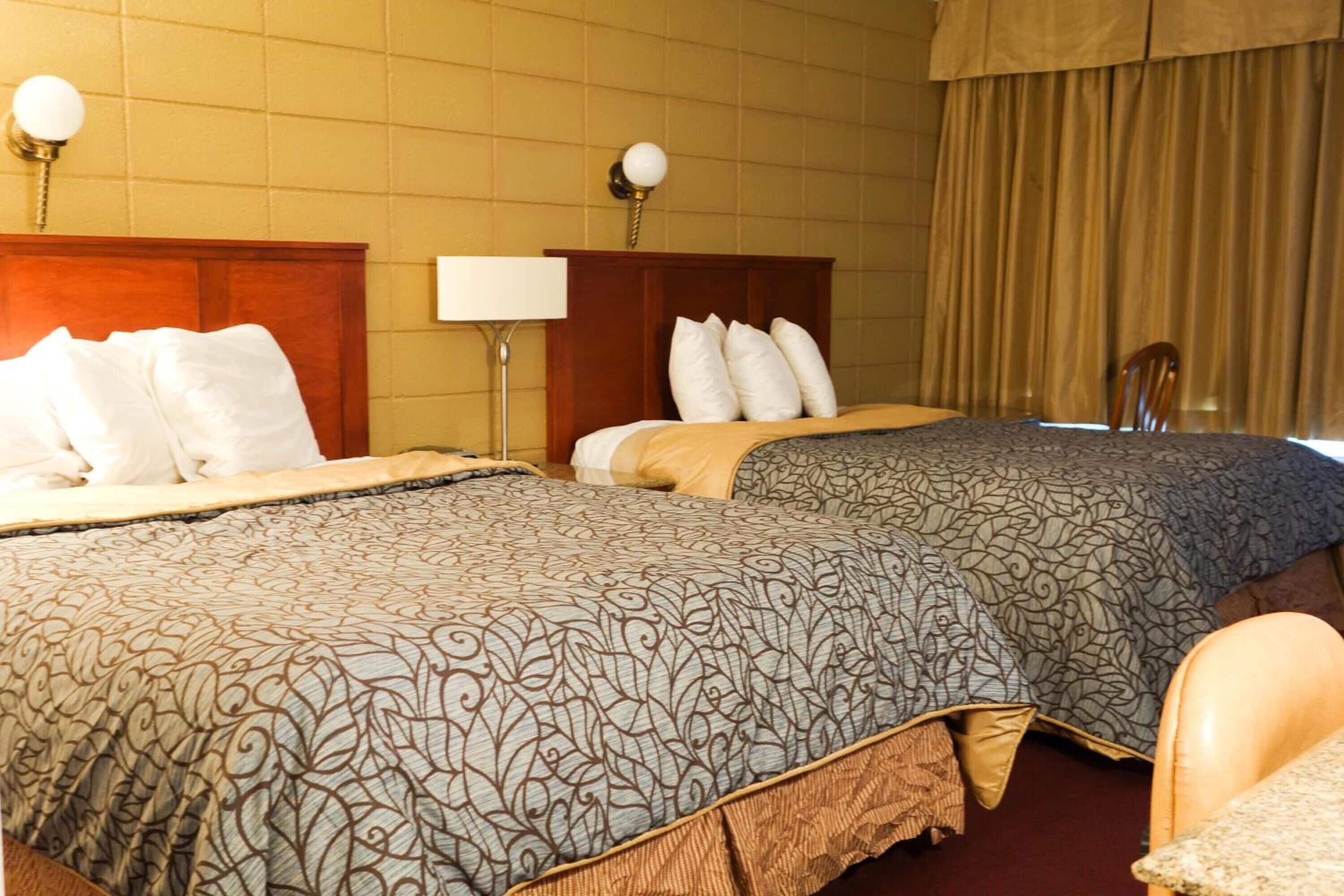Ein Hotelzimmer mit zwei Betten, einem Holz-Kopfteil, einer Nachttischlampe, einem Fenster mit Vorhängen und einem Sessel.