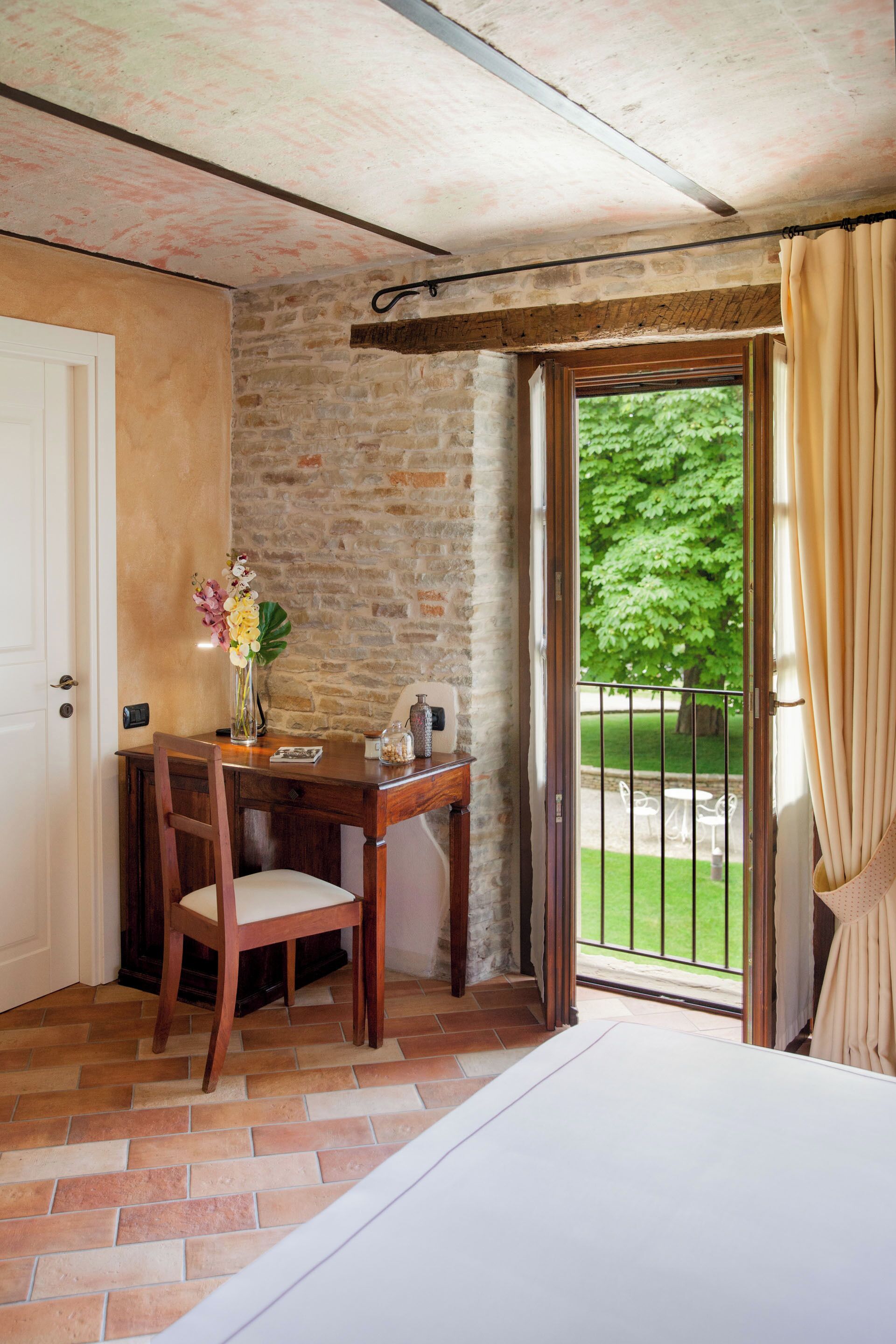 A room with a wooden desk, chair, and a vase of flowers. There is a door, a window with curtains, and a view of a green outdoor area.