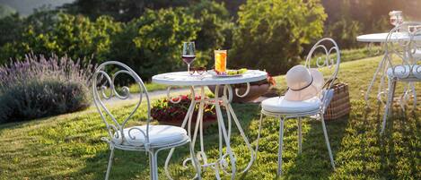 Un jardín con una mesa redonda y sillas, una copa de vino tinto, un vaso alto de zumo de naranja y una cesta de fruta.