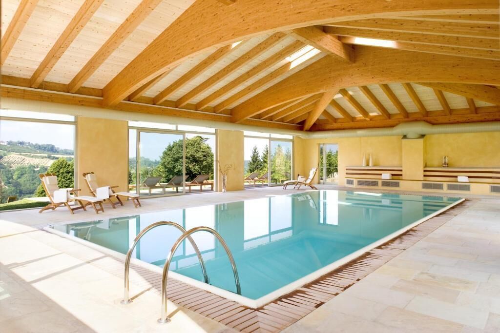 An indoor swimming pool with wooden beams, clear glass walls, and sunlit areas with lounge chairs.