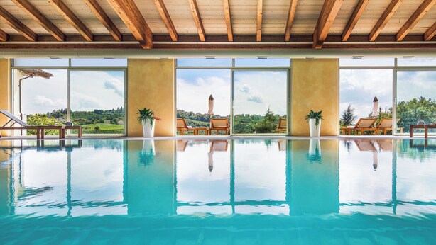 A large indoor swimming pool with clear blue water, wooden ceiling beams, and large windows offering views of greenery and hills. - Hotel Relais Montemarino (Borgomale)
