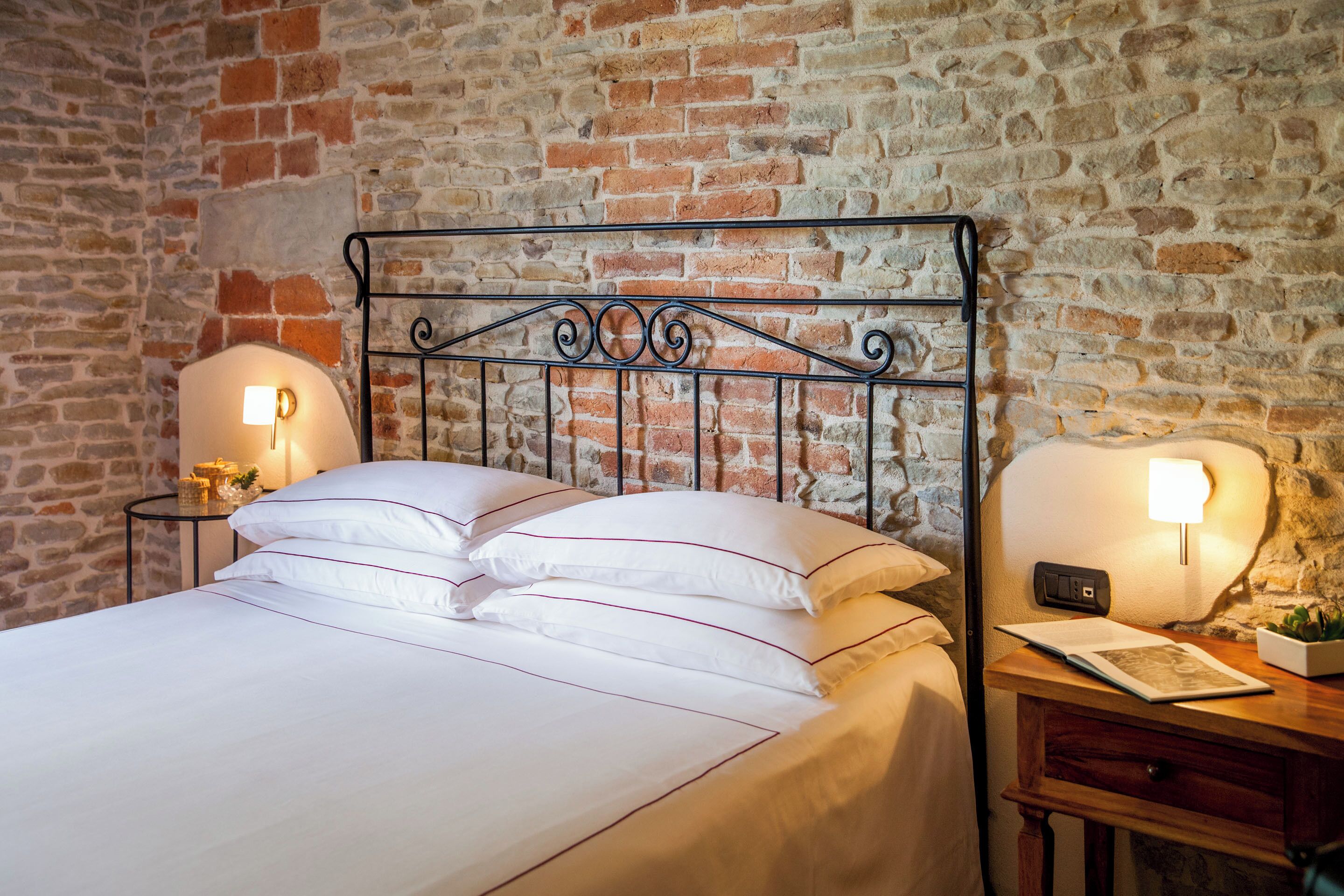 Una cama bien hecha con sábanas blancas y ribetes rojos, flanqueada por dos mesitas de noche con lámparas, frente a una pared de ladrillo.