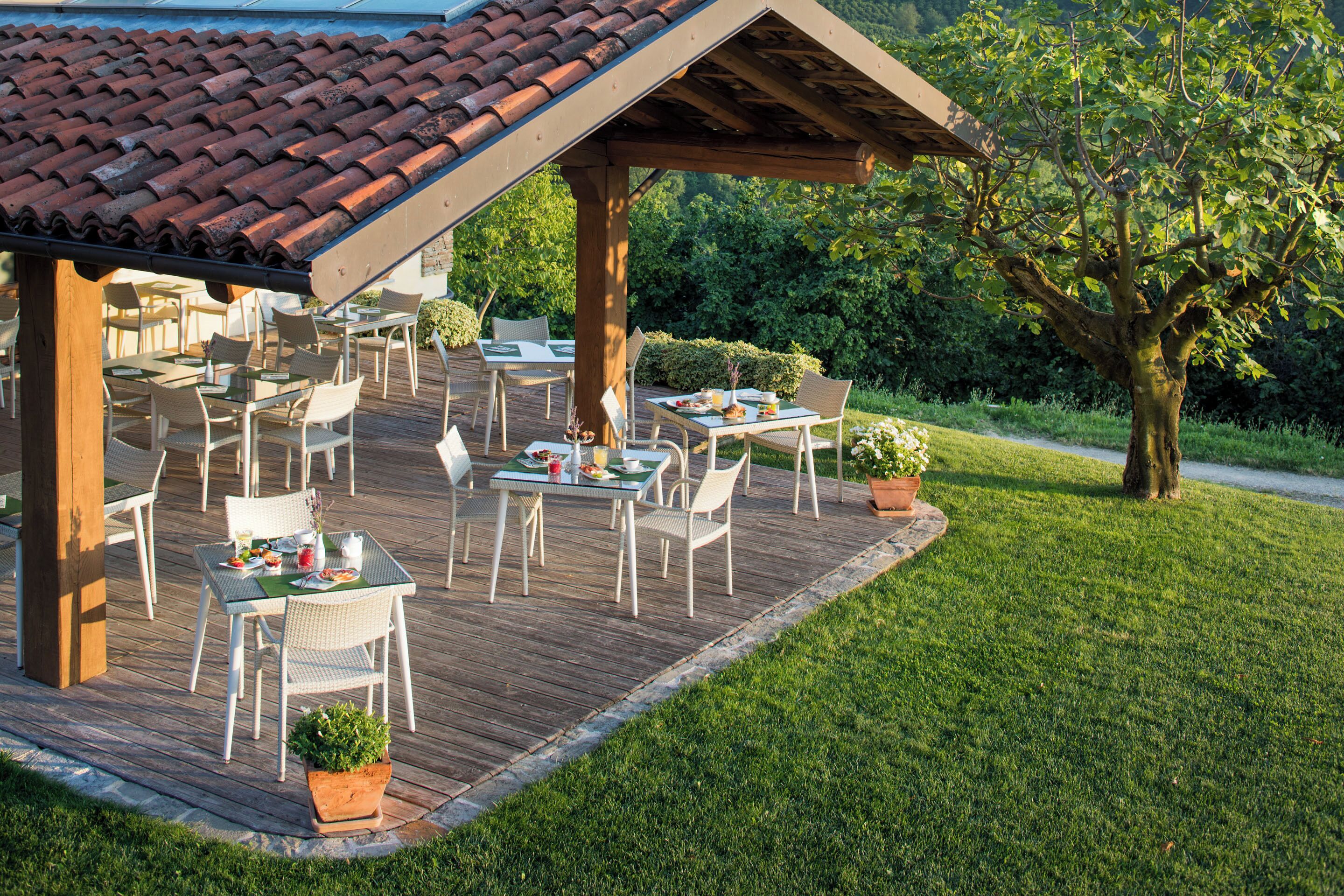 An outdoor patio with tables and chairs, surrounded by greenery and a wooden deck.
