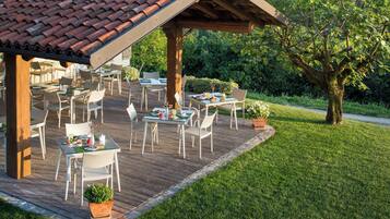 An outdoor patio with tables and chairs, surrounded by greenery and a wooden deck.