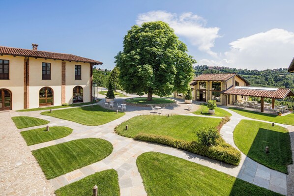 A courtyard with a large tree, stone pathways, and outdoor seating areas. - Hotel Relais Montemarino (Borgomale)