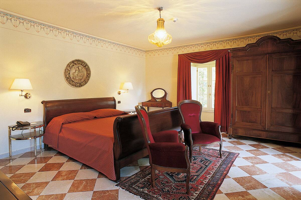 A hotel room with a bed, a chair, a desk, a lamp, a wardrobe, and a patterned rug.
