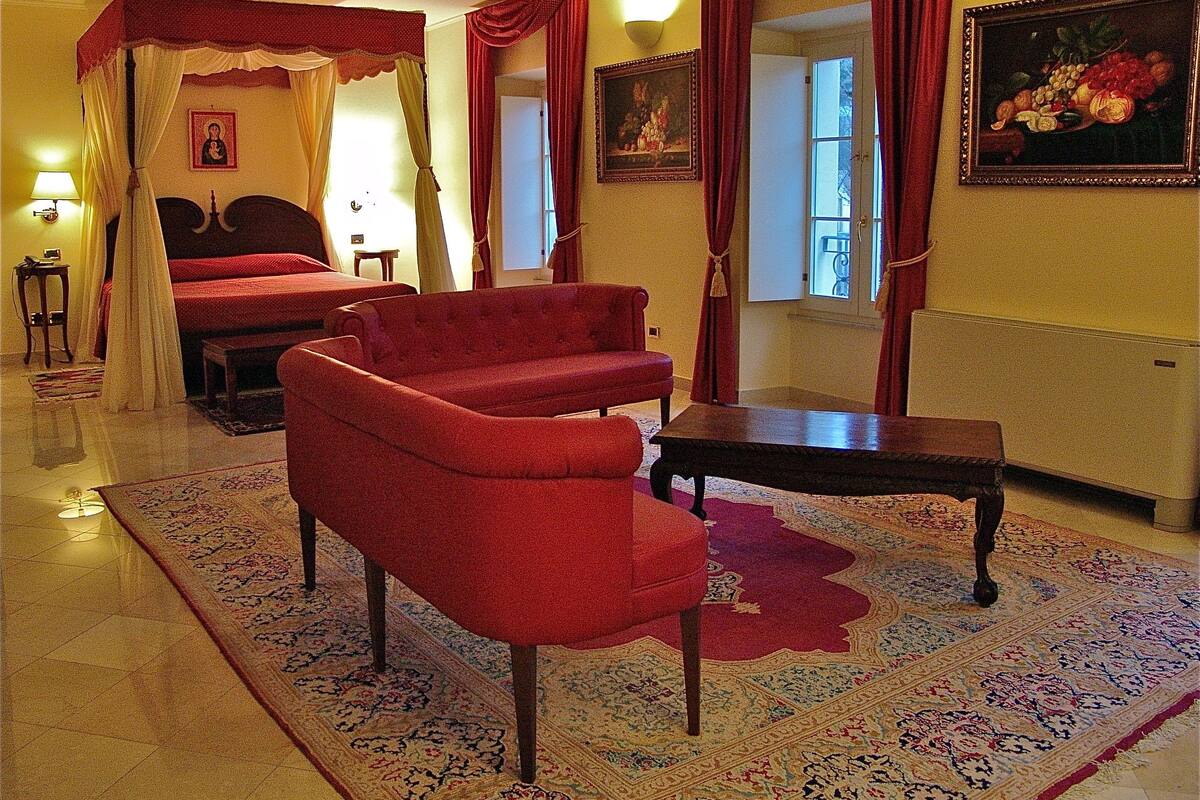 A hotel room with a four-poster bed, a red sofa, a small table, and a large window with curtains.