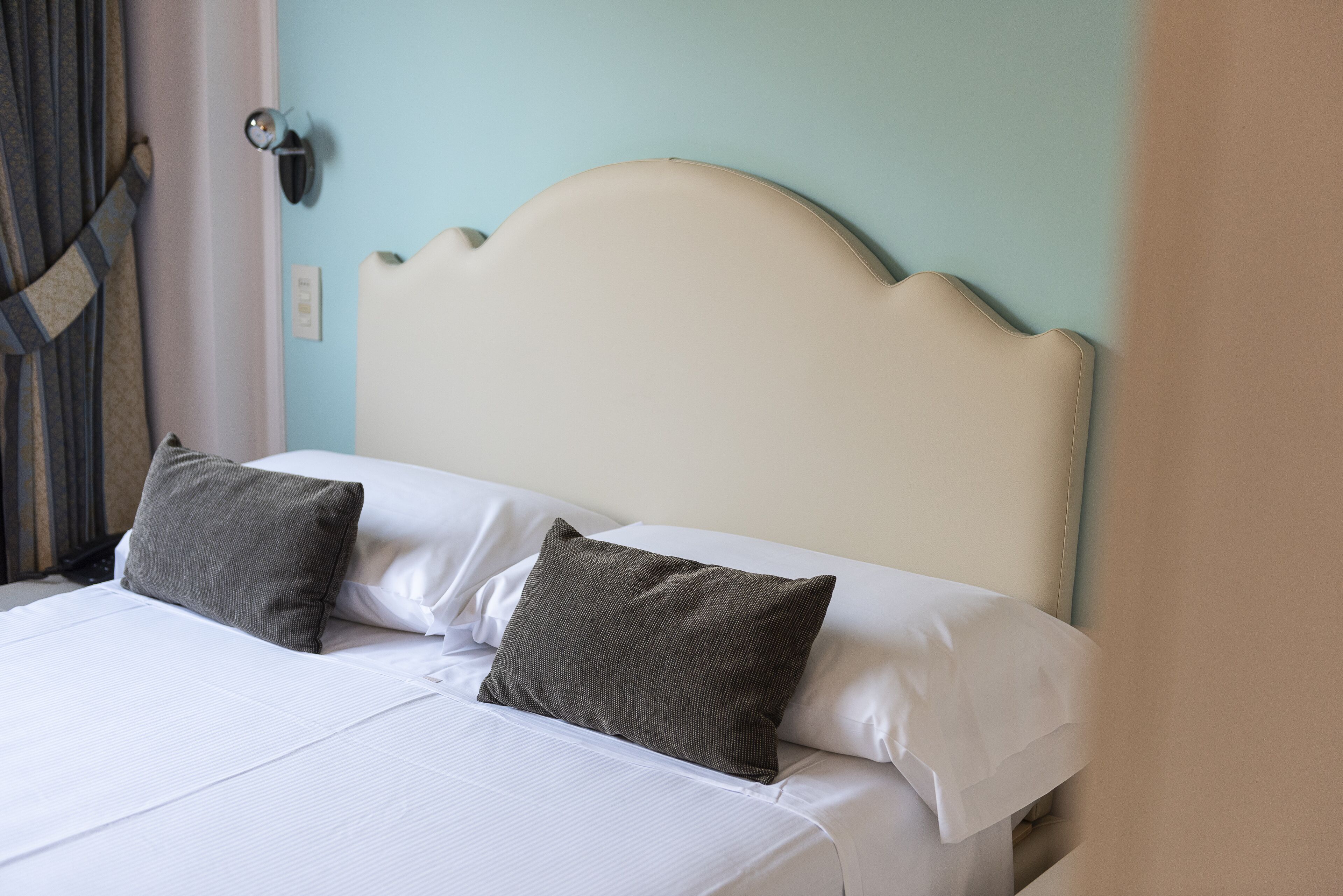 Un lit avec du linge de lit blanc et deux oreillers gris foncé, une tête de lit de couleur claire et un luminaire fixé au mur.