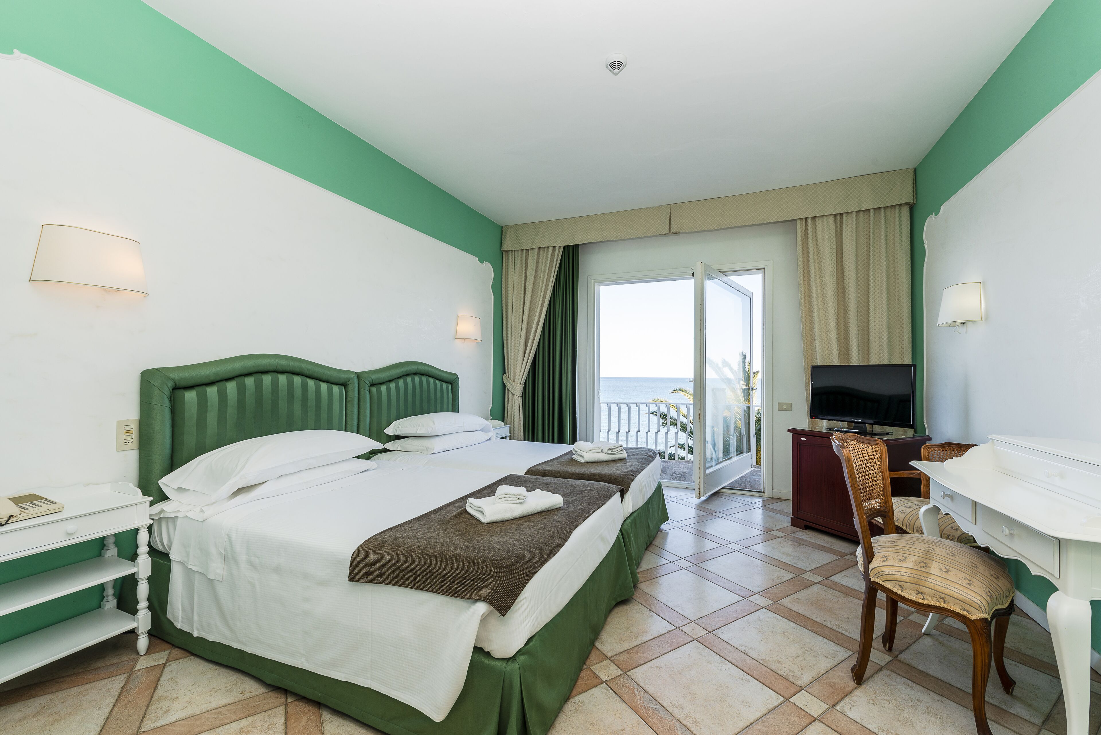 Una camera d'albergo con un letto, una scrivania, una sedia, una televisione e un balcone con tende.