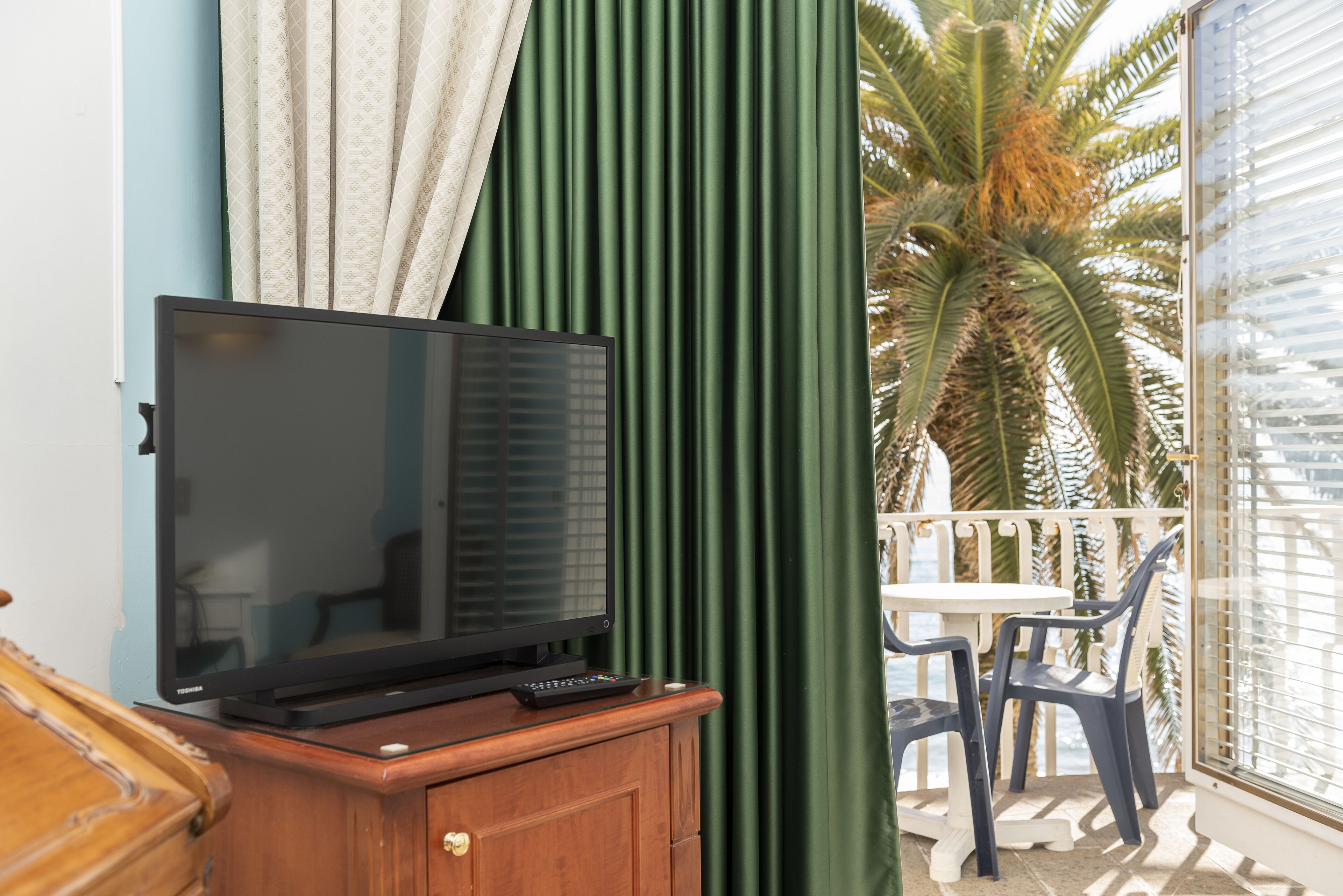 Una televisione su un mobile in legno, un tavolo con sedie e vista su una palma attraverso una finestra.