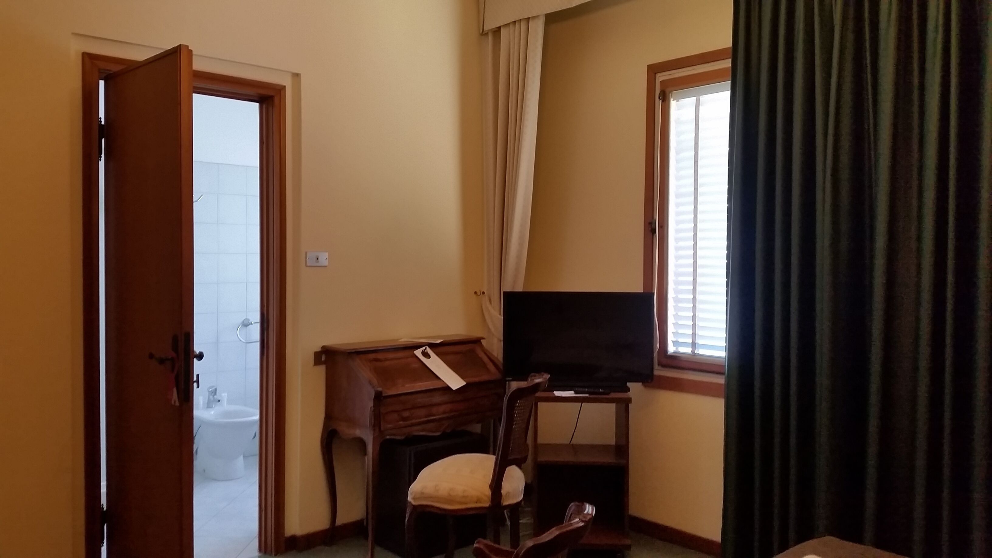 A small room with a wooden desk, a chair, a television, a window with curtains, and a door leading to a bathroom.