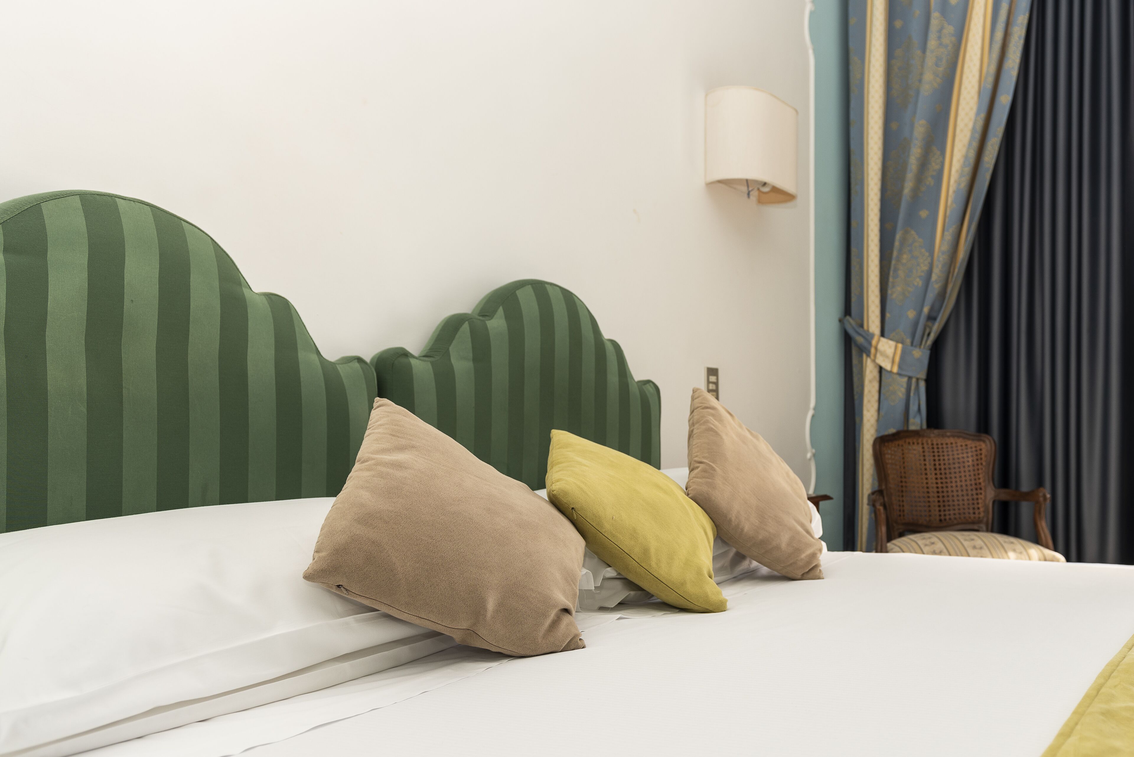 Un lit avec une tête de lit à rayures, des oreillers beige et jaunes, un matelas blanc et une chaise en bois, dans une pièce dotée d’une lampe fixée au mur et de rideaux.