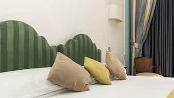 Un lit avec une tête de lit à rayures, des oreillers beige et jaunes, un matelas blanc et une chaise en bois, dans une pièce dotée d’une lampe fixée au mur et de rideaux.