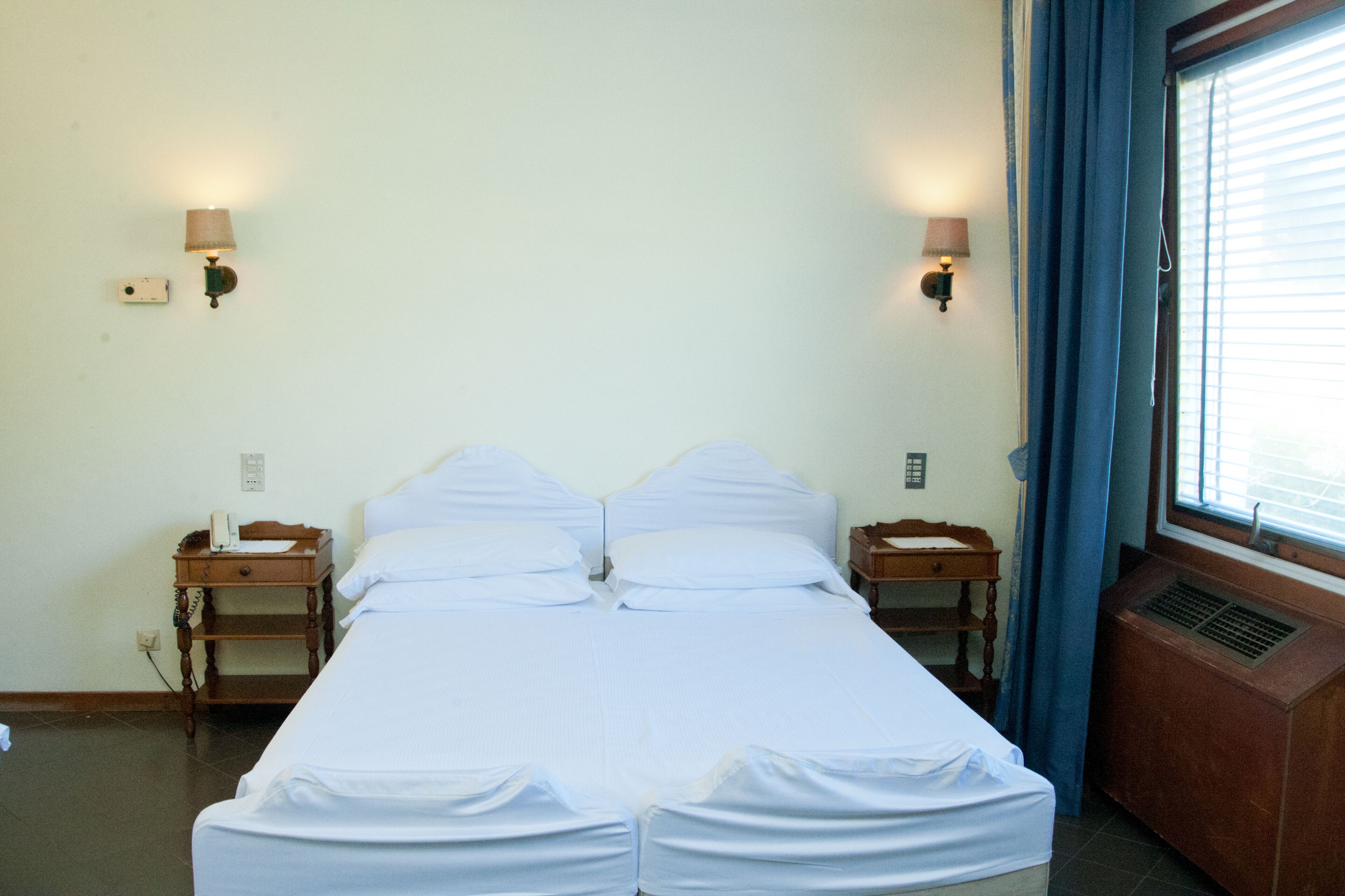 Una cama doble con ropa de cama blanca, dos mesitas de noche con lámparas, una ventana con persianas y un aire acondicionado de pared.