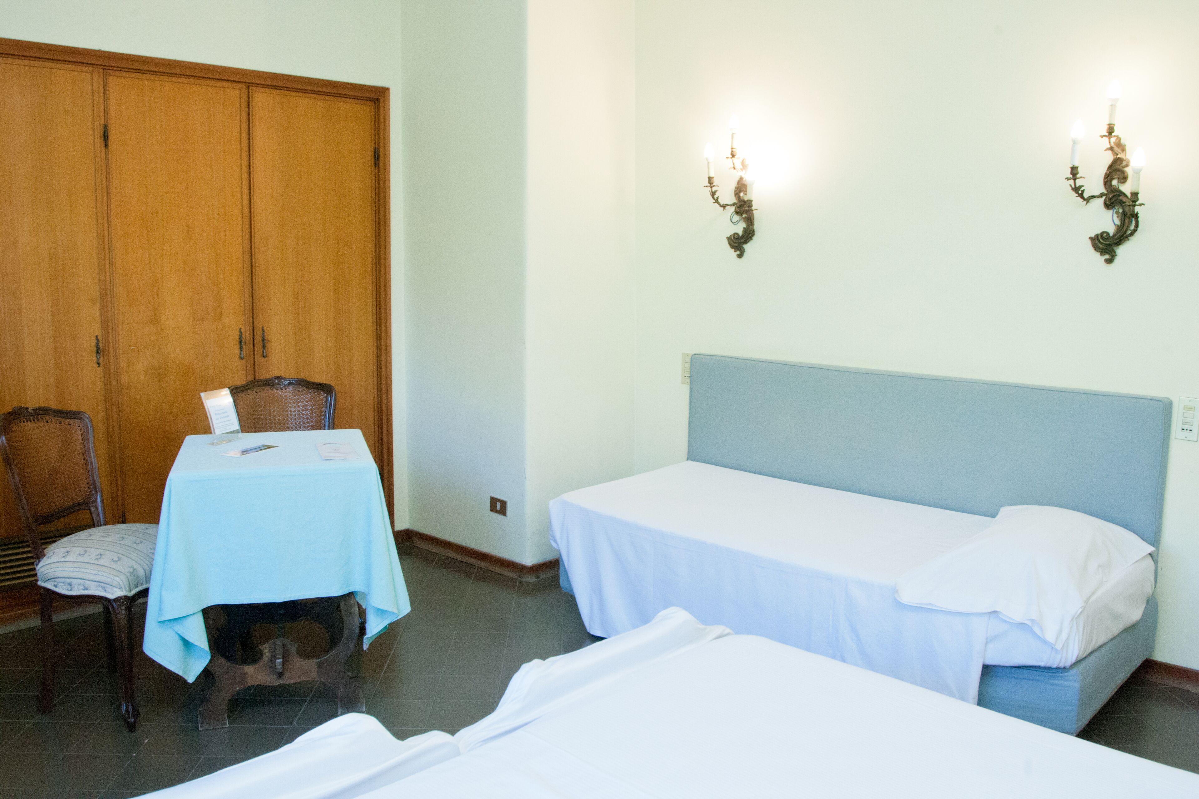 Une chambre avec deux lits simples, une armoire en bois, une table recouverte d’une nappe bleue et deux appliques murales.