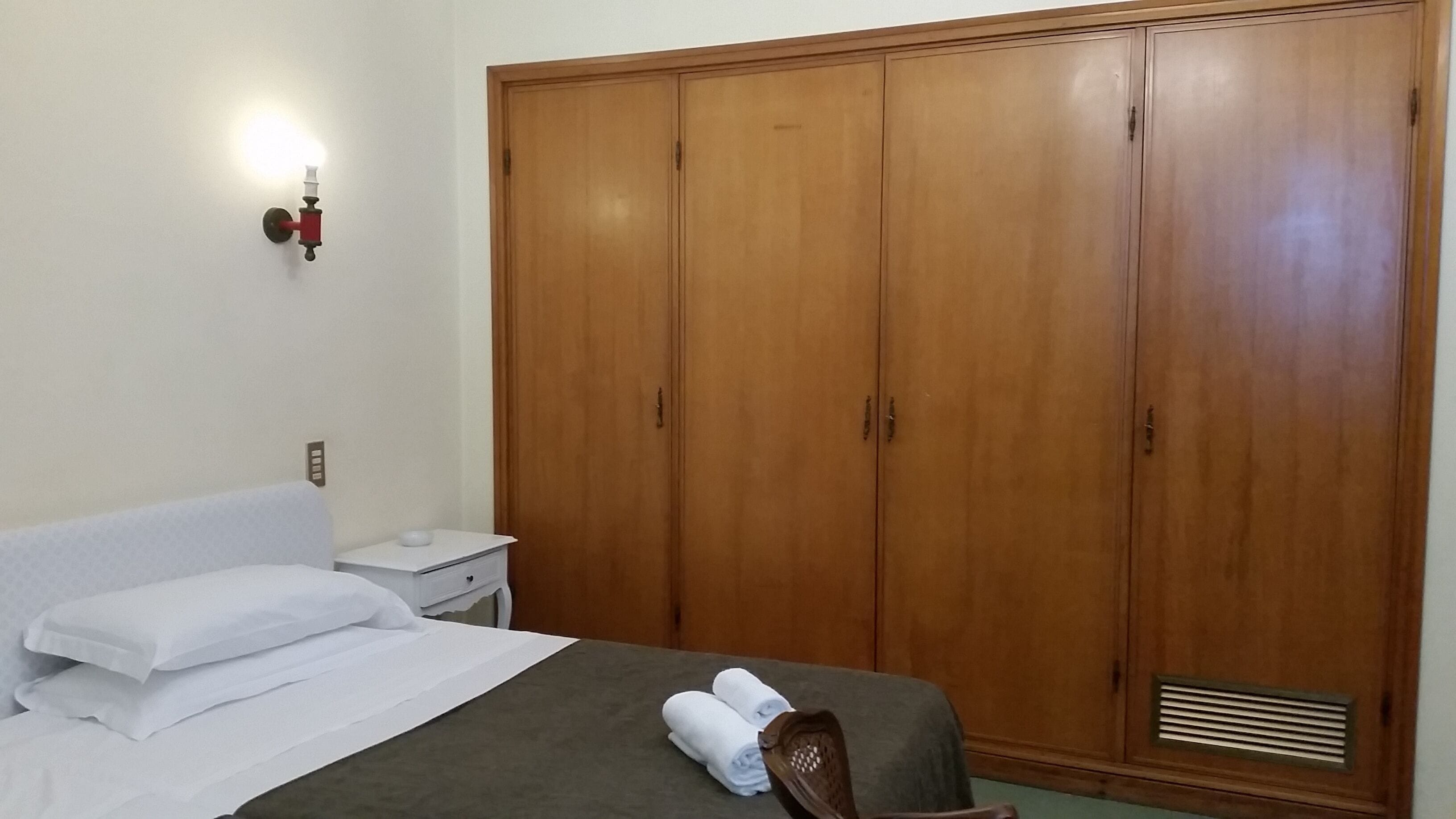 Une chambre d’hôtel avec un lit, des oreillers blancs, une table de chevet, une lampe et une grande armoire en bois.