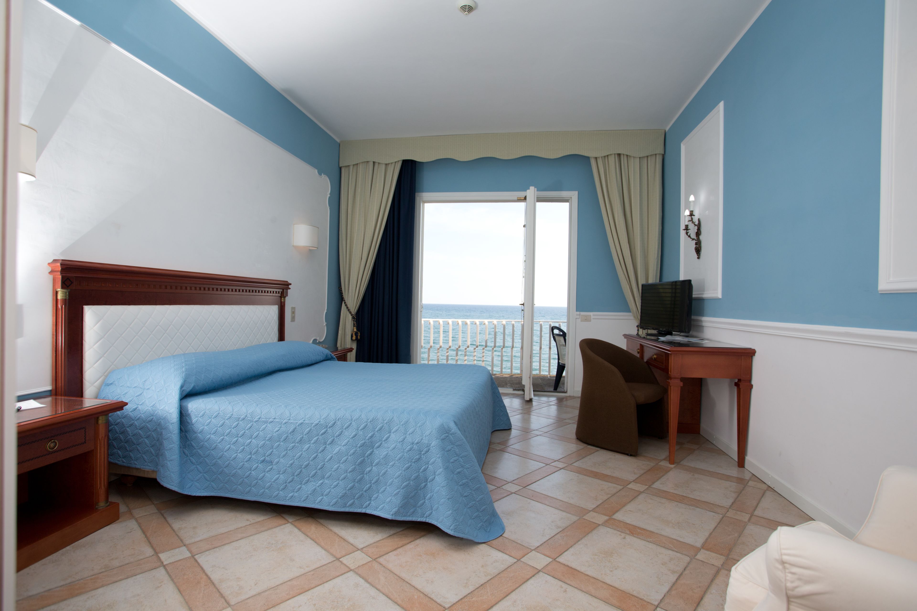 Habitación de hotel con cama, escritorio, silla, televisor y vista al mar a través de una ventana.