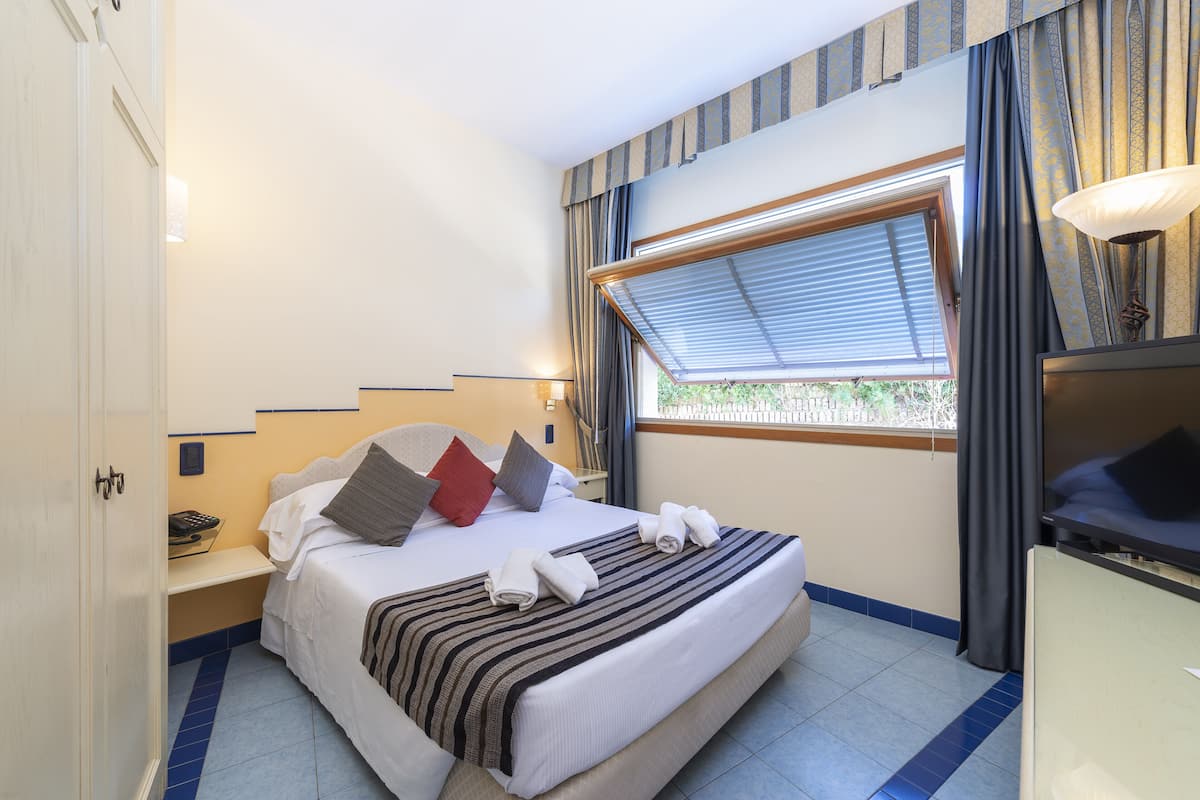 Ein Hotelzimmer mit Bett, Nachttisch, einem Fenster mit Vorhängen und Blick auf Grün.