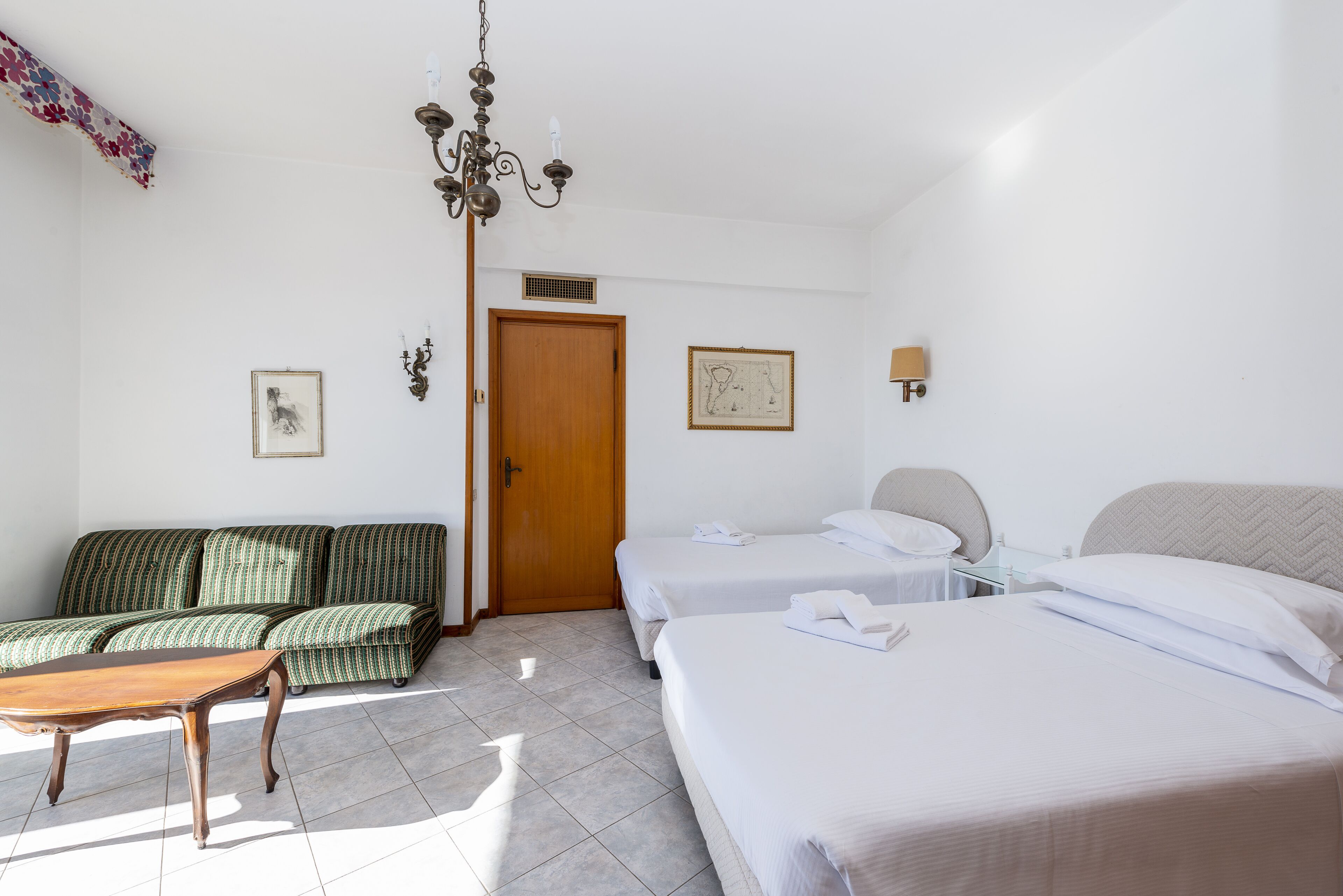 A hotel room with a sofa set, two single beds, a wooden door, and a chandelier.
