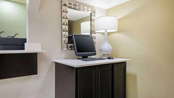A hotel room with a desk, computer, lamp, and a mirror with a geometric frame.