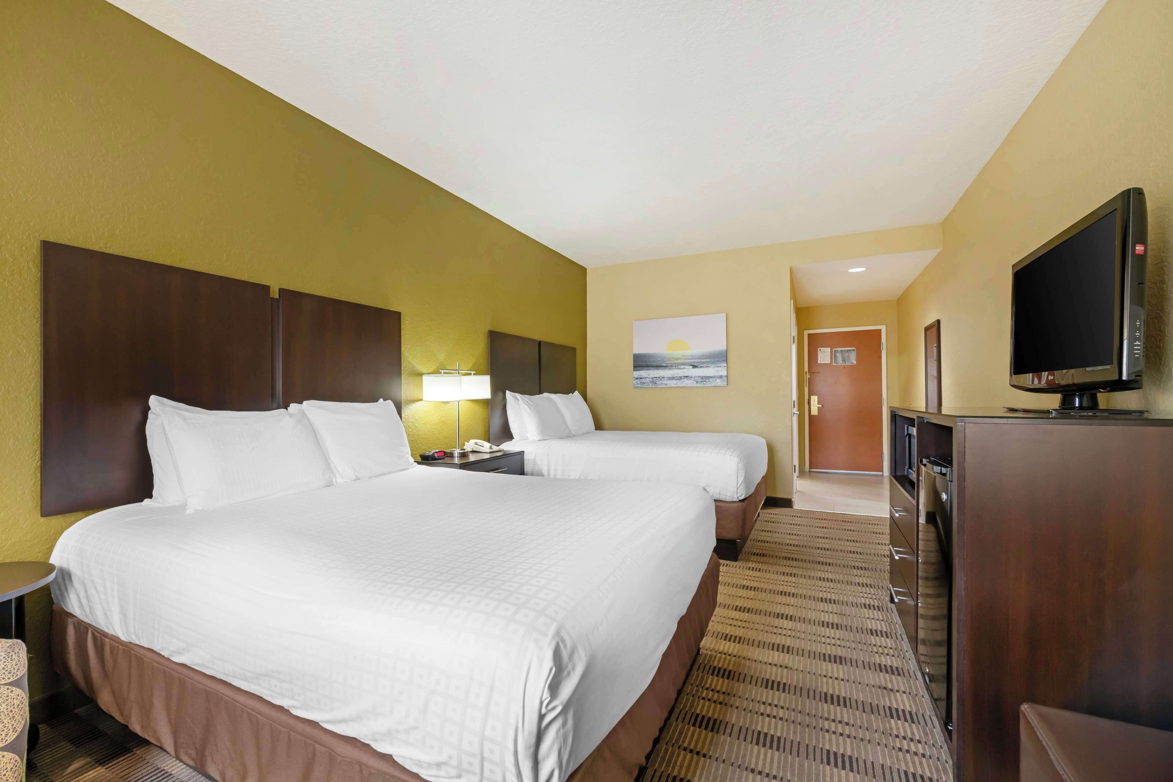 A hotel room with two beds, a television, a desk with a lamp, and a painting of a sunset.