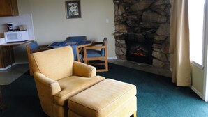 A living room with a stone fireplace, a dining table with chairs, a kitchen area with a microwave, and a large window with curtains. - Ocean Shores Inn & Suites (Ocean Shores)